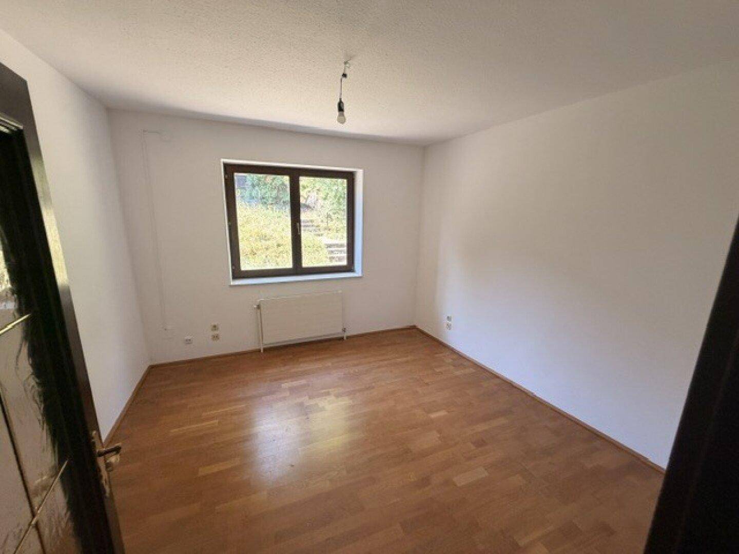 Leeres Zimmer mit Holzfußboden, weißen Wänden und einem Fenster mit Blick ins Grüne.