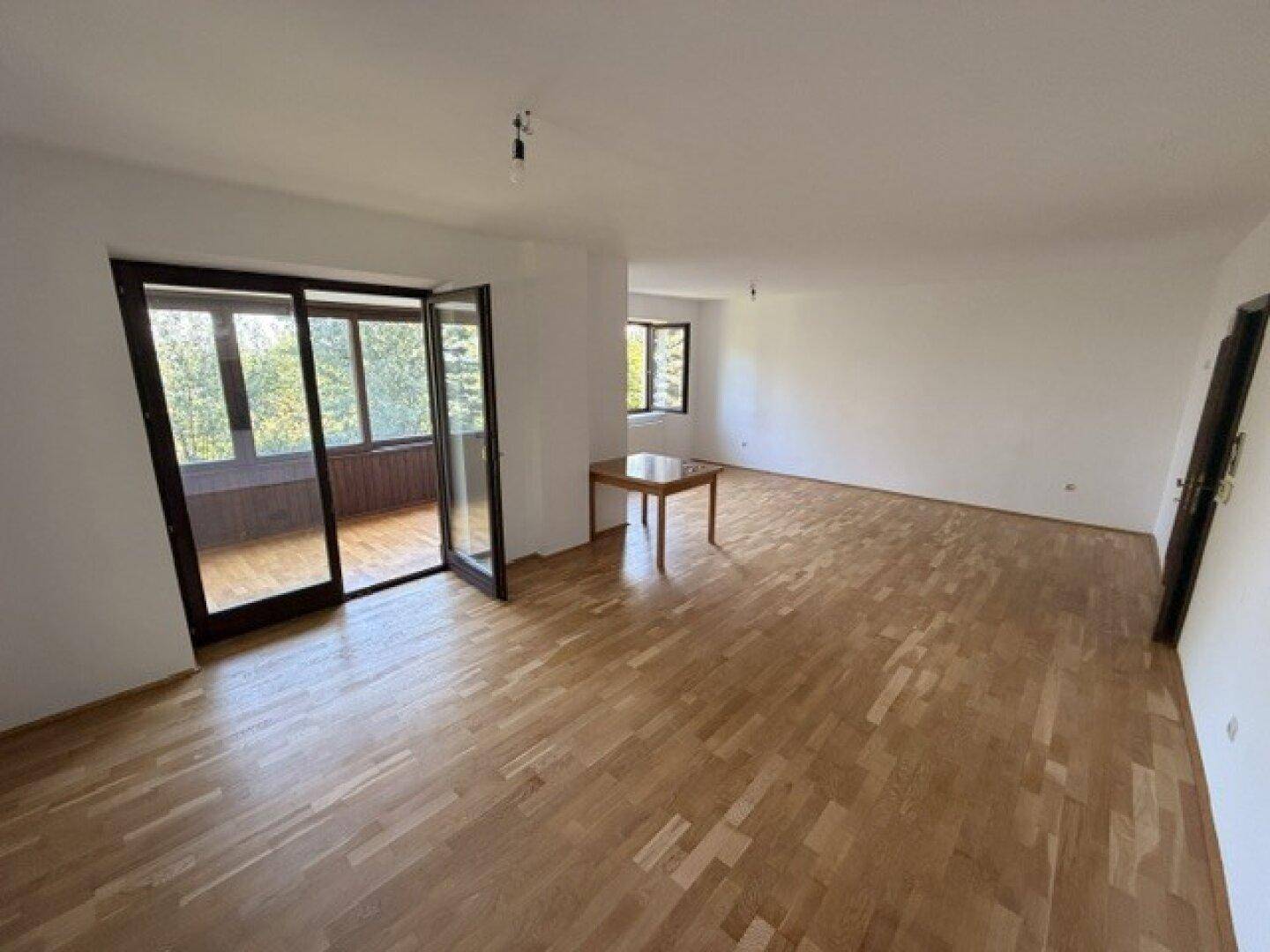 Geräumiges Wohnzimmer mit Holzfußboden, weißen Wänden und Zugang zu einem verglasten Balkon.