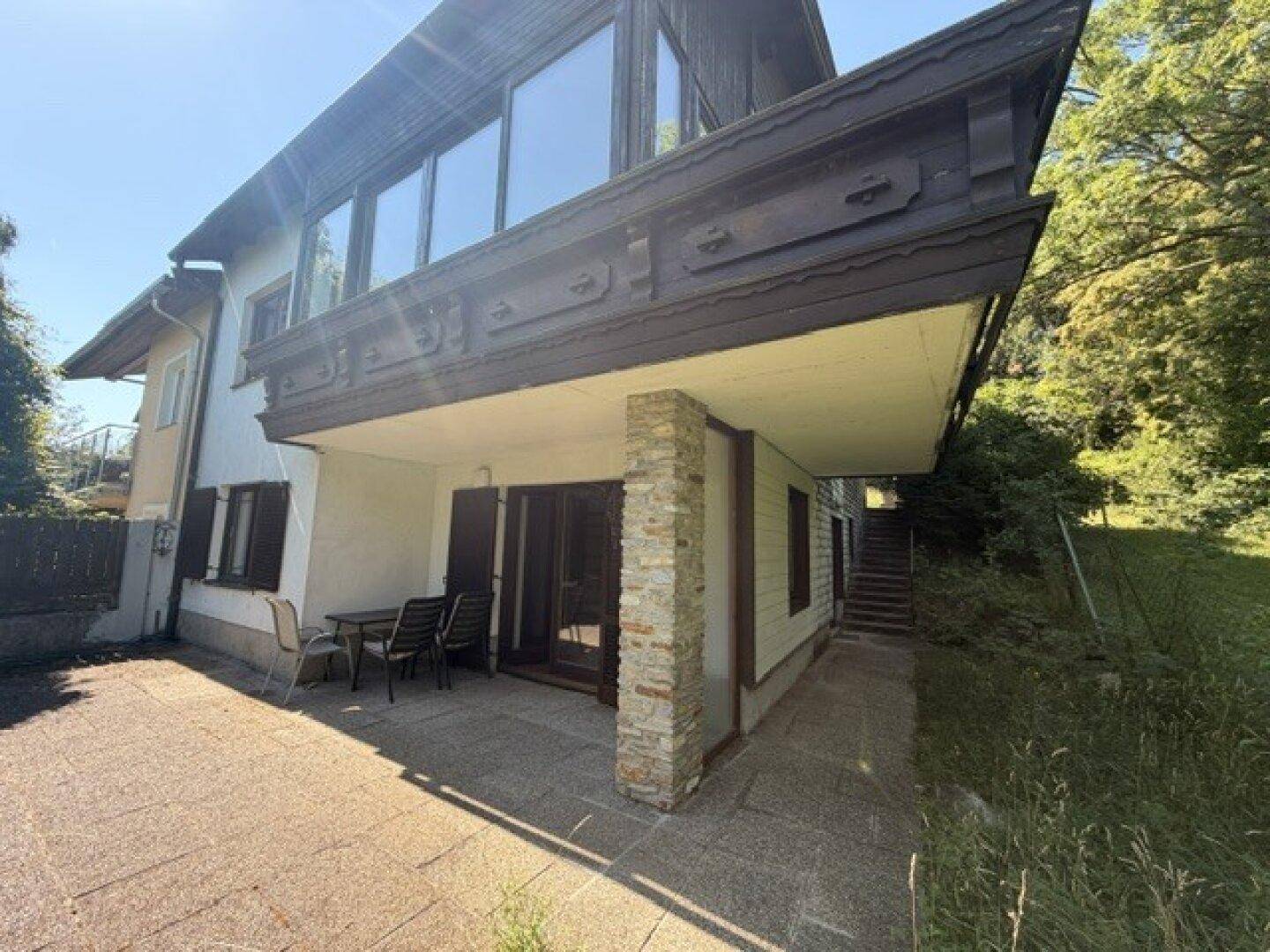 Hausfassade mit großer gepflasterter Terrasse, Balkon und Treppe, die zum oberen Bereich führt.