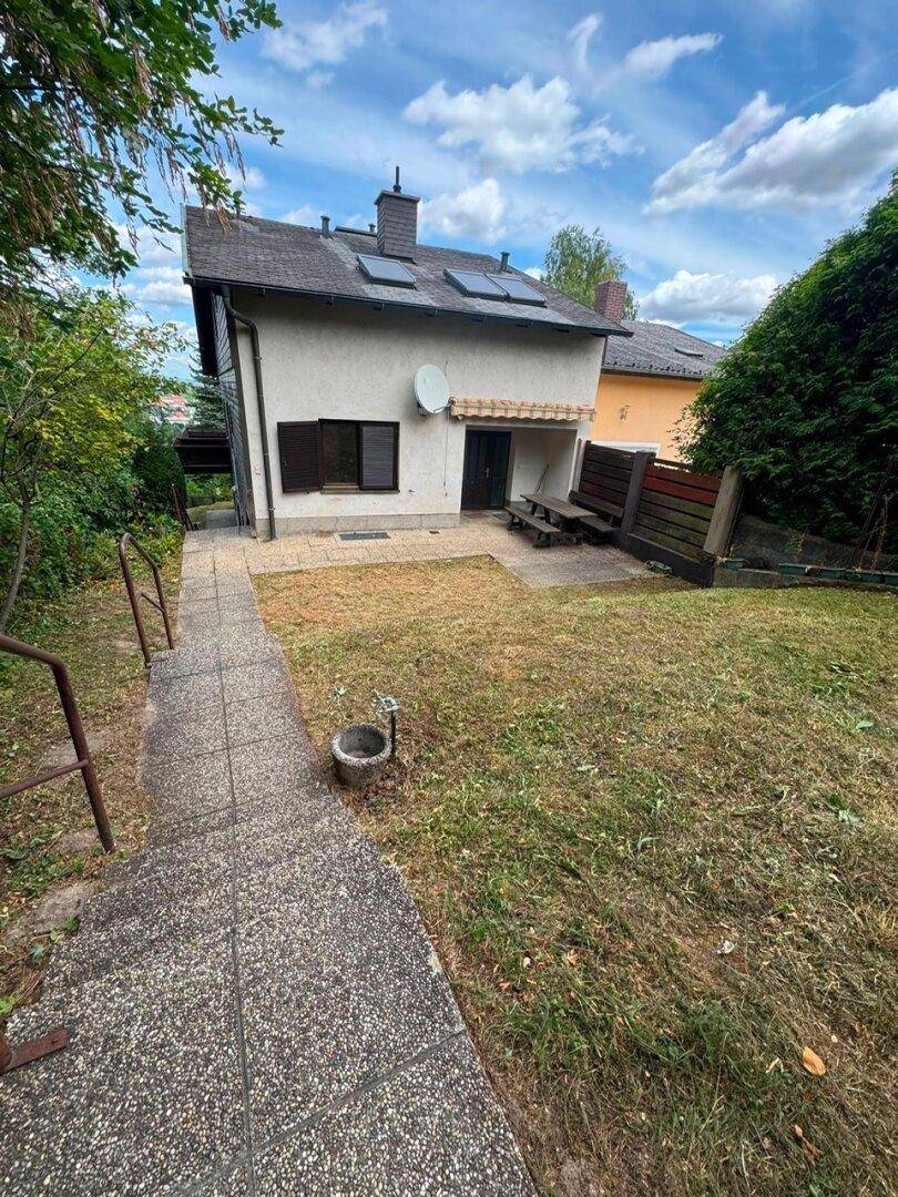 Hinterhof des Hauses mit gepflastertem Bereich, Rasenfläche und einem Weg, der den Hang hinaufführt.