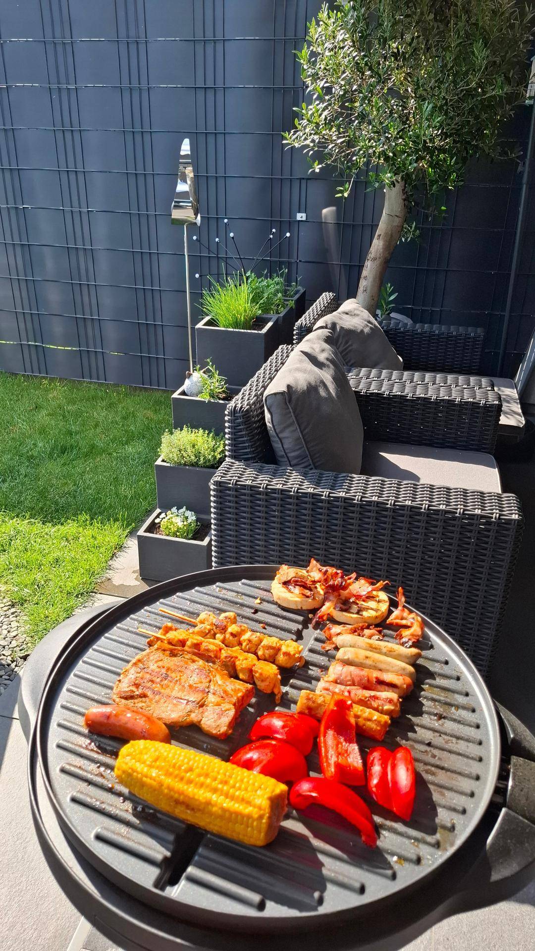 Grillbereich im Garten mit leckerem Essen auf dem Grill und bequemen Sitzgelegenheiten im Hintergrund.