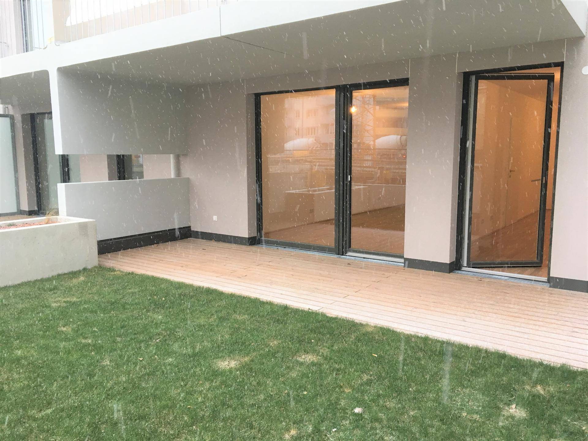 Geräumige Terrasse mit Holzdeck und angrenzender Rasenfläche, zugänglich durch große Glastüren des Neubaus.