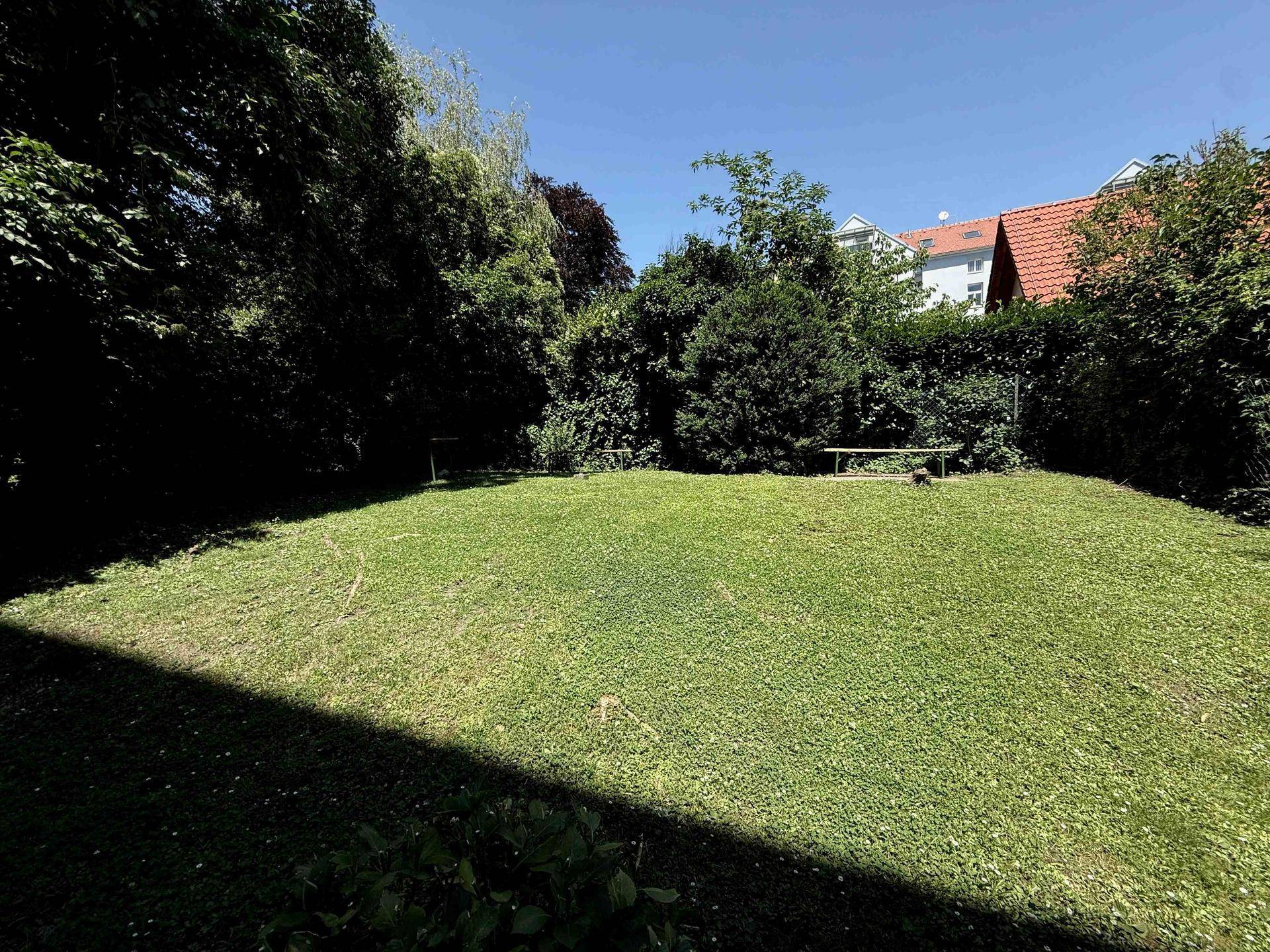 Weitläufige Rasenfläche im Gemeinschaftsgarten mit schattenspendenden Bäumen und einer Sitzbank im Hintergrund.