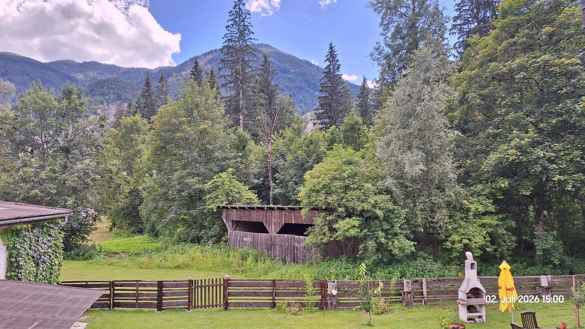 Weitläufiger Gartenbereich mit Grillplatz, umgeben von hohen Bäumen und einer beeindruckenden Bergkulisse.
