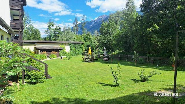 Großer, gepflegter Rasenbereich mit Gartenmöbeln und Grillplatz, umgeben von Bäumen und mit Bergblick.