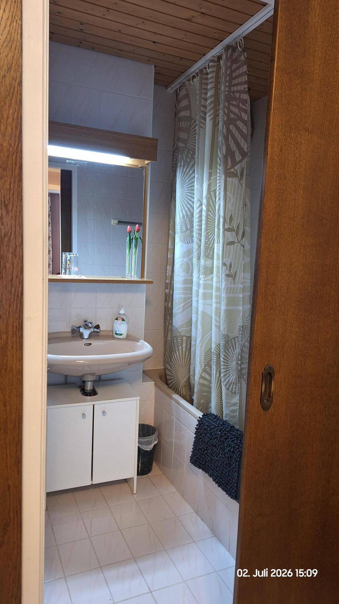 Badezimmer mit Badewanne, Duschvorhang, Waschbecken und Spiegelschrank unter einer Holzdecke.