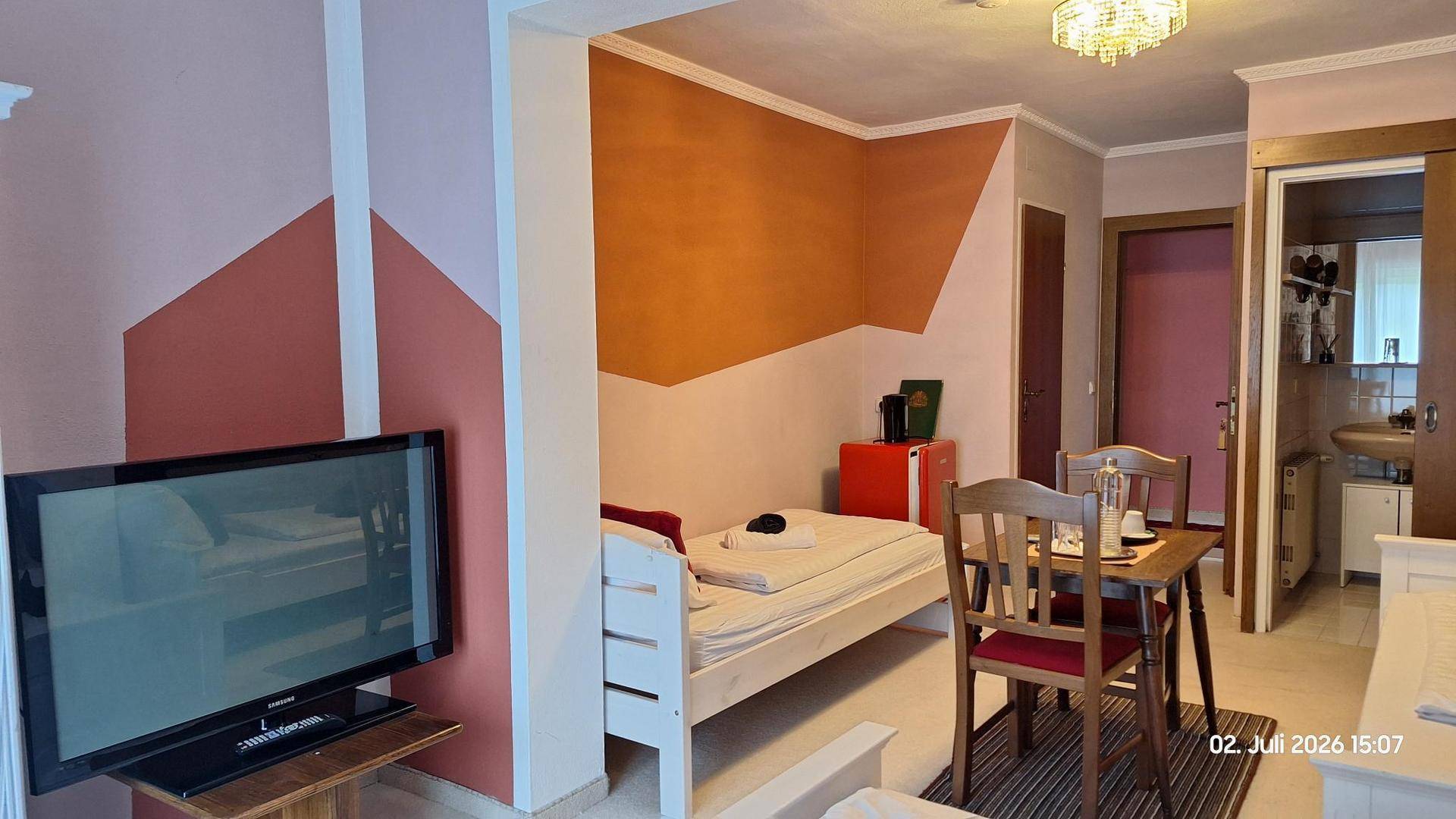 Schlafzimmer mit Einzelbett, Fernseher, Minikühlschrank und einem Tisch mit Stühlen, mit farbigen Wandakzenten.
