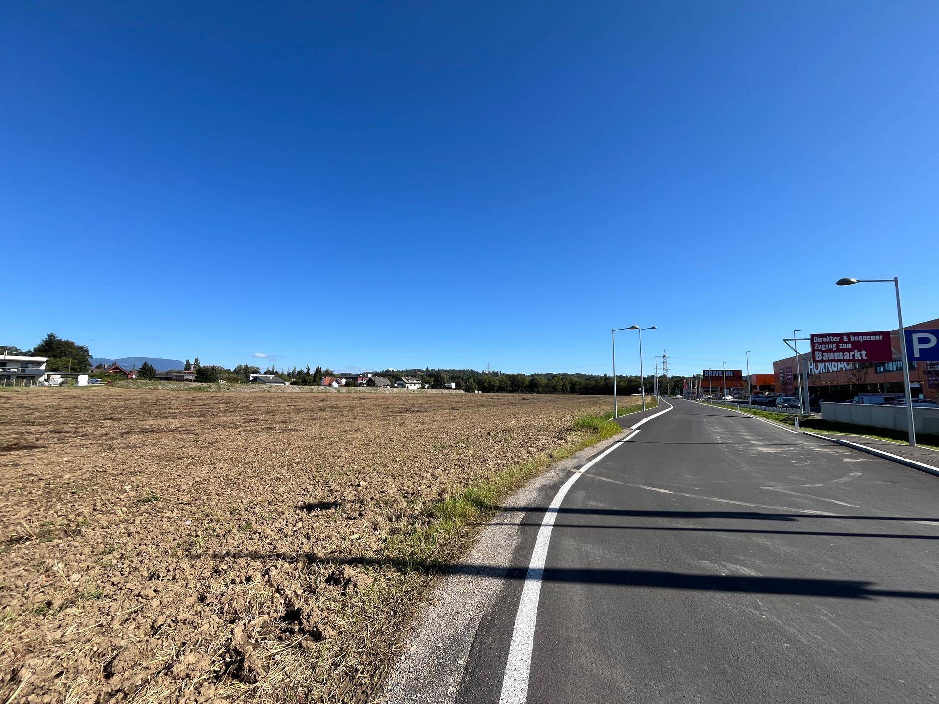 Weitläufiges Feld neben einer neuen Straße, mit Gewerbegebäuden und einem Baumarkt in der Ferne unter blauem Himmel.