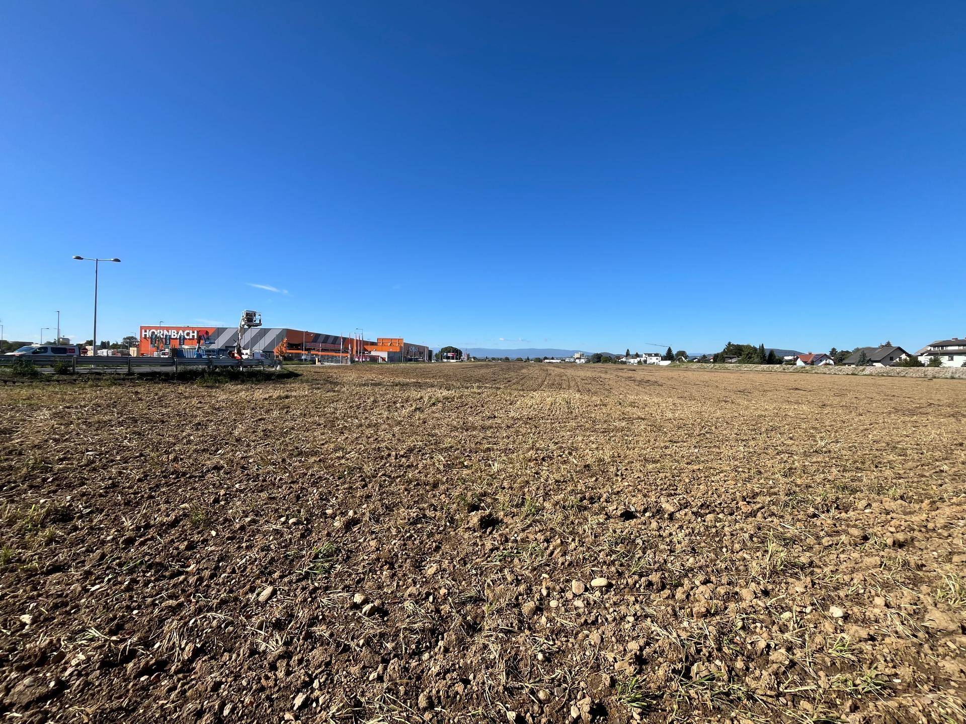 Weitläufiges, frisch gepflügtes Feld mit einem großen Baumarktgebäude und anderen Gewerbebauten im Hintergrund.