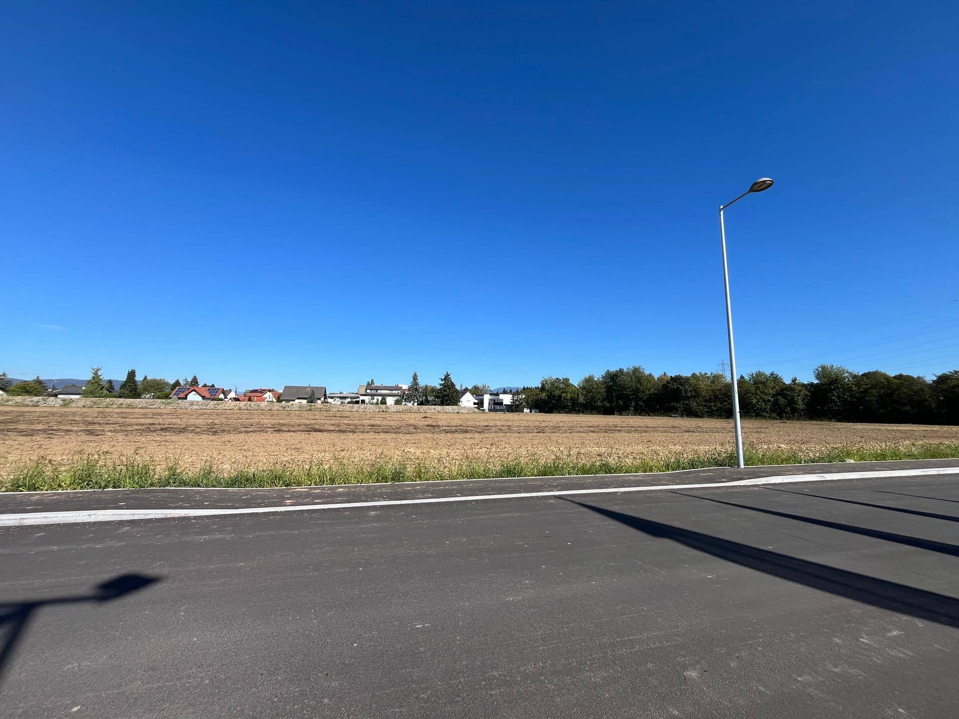 Weitläufiges Feld grenzt an eine neue Straße, mit einer Siedlung und Waldrand in der Ferne unter klarem Himmel.