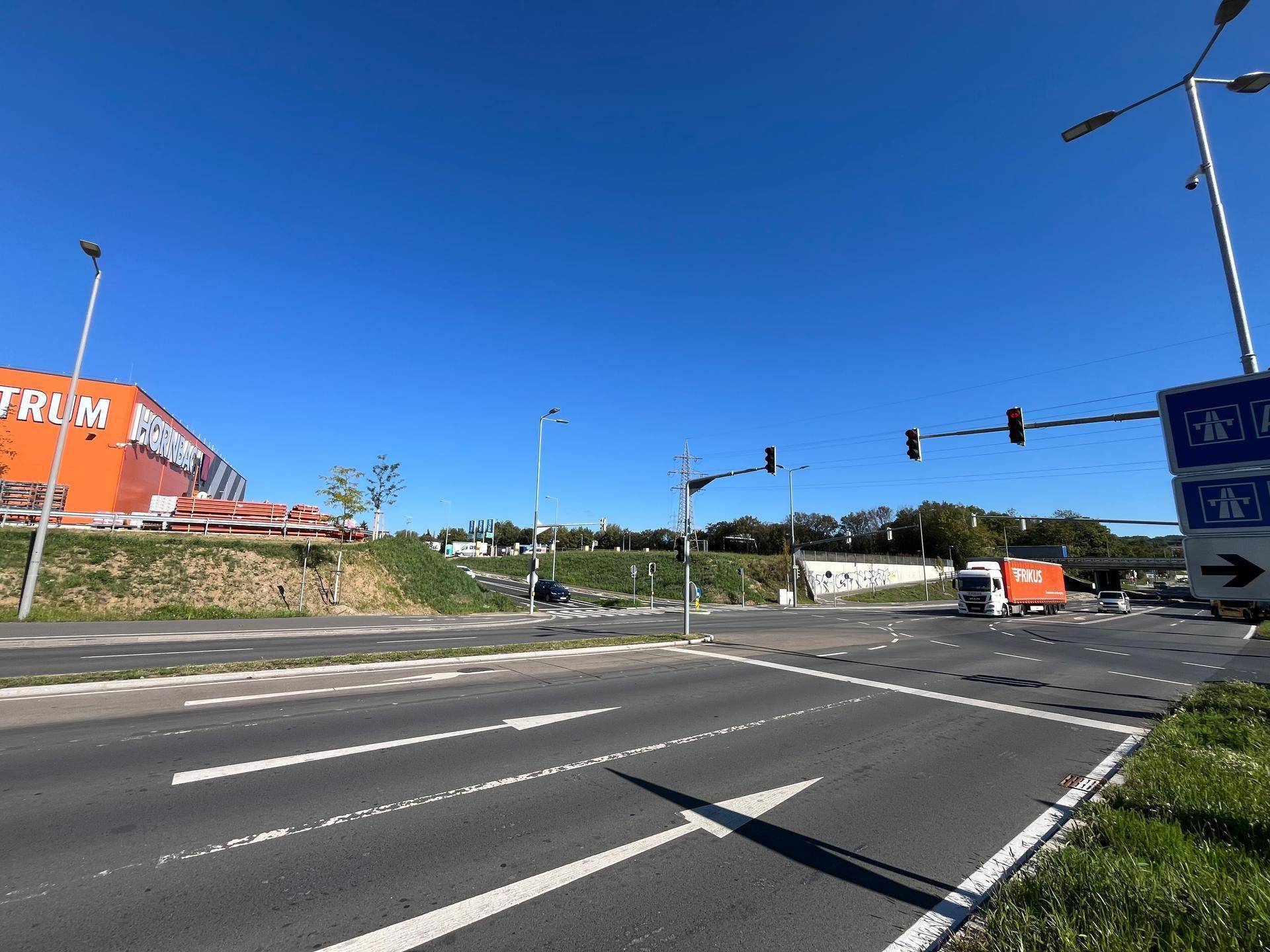 Verkehrskreuzung mit Ampeln und einem großen Baumarktgebäude, daneben eine Autobahnauffahrt unter blauem Himmel.
