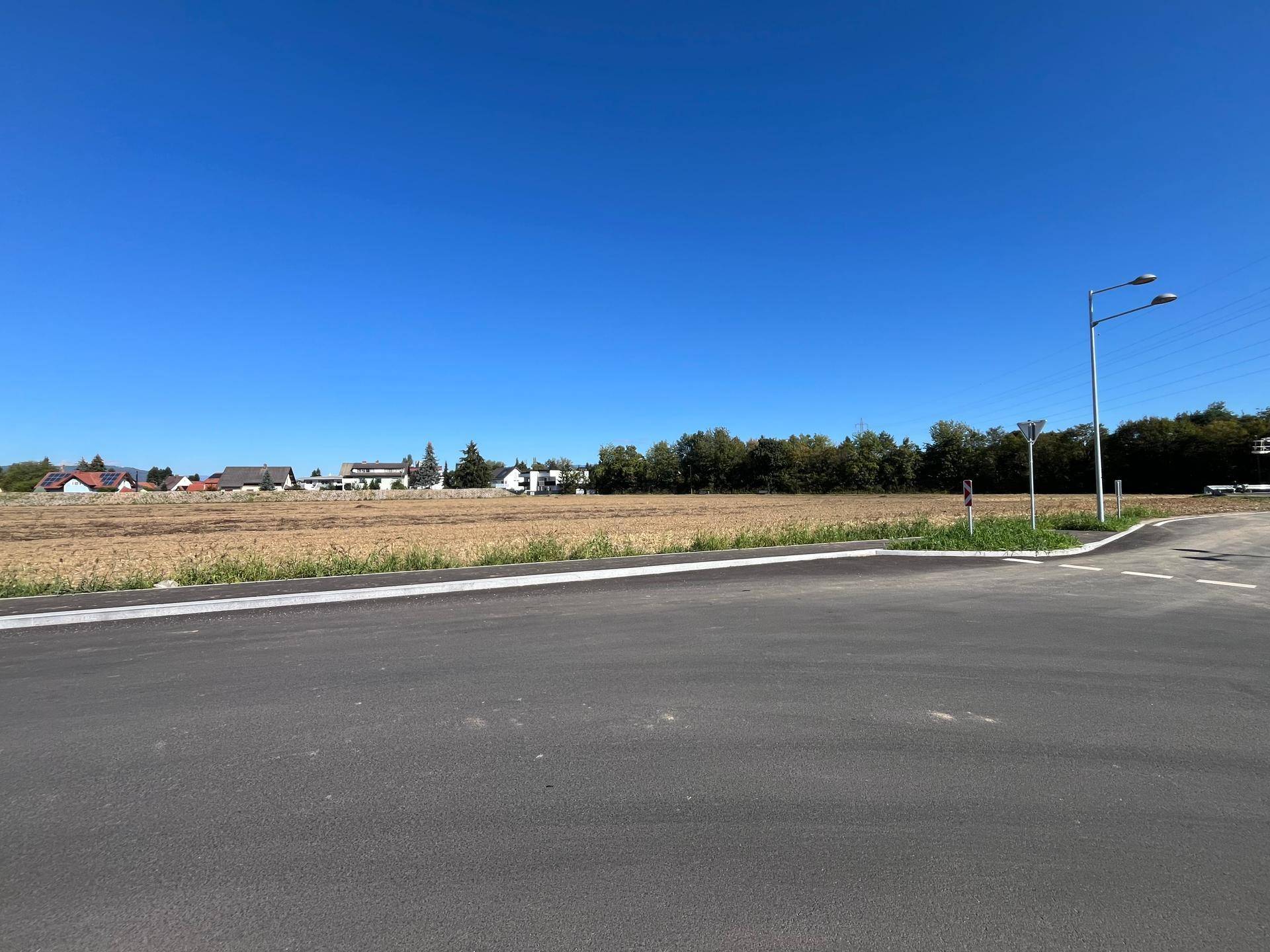 Neue Straßenkreuzung neben einem weiten Feld, mit Wohnhäusern und Bäumen am Horizont unter strahlend blauem Himmel.