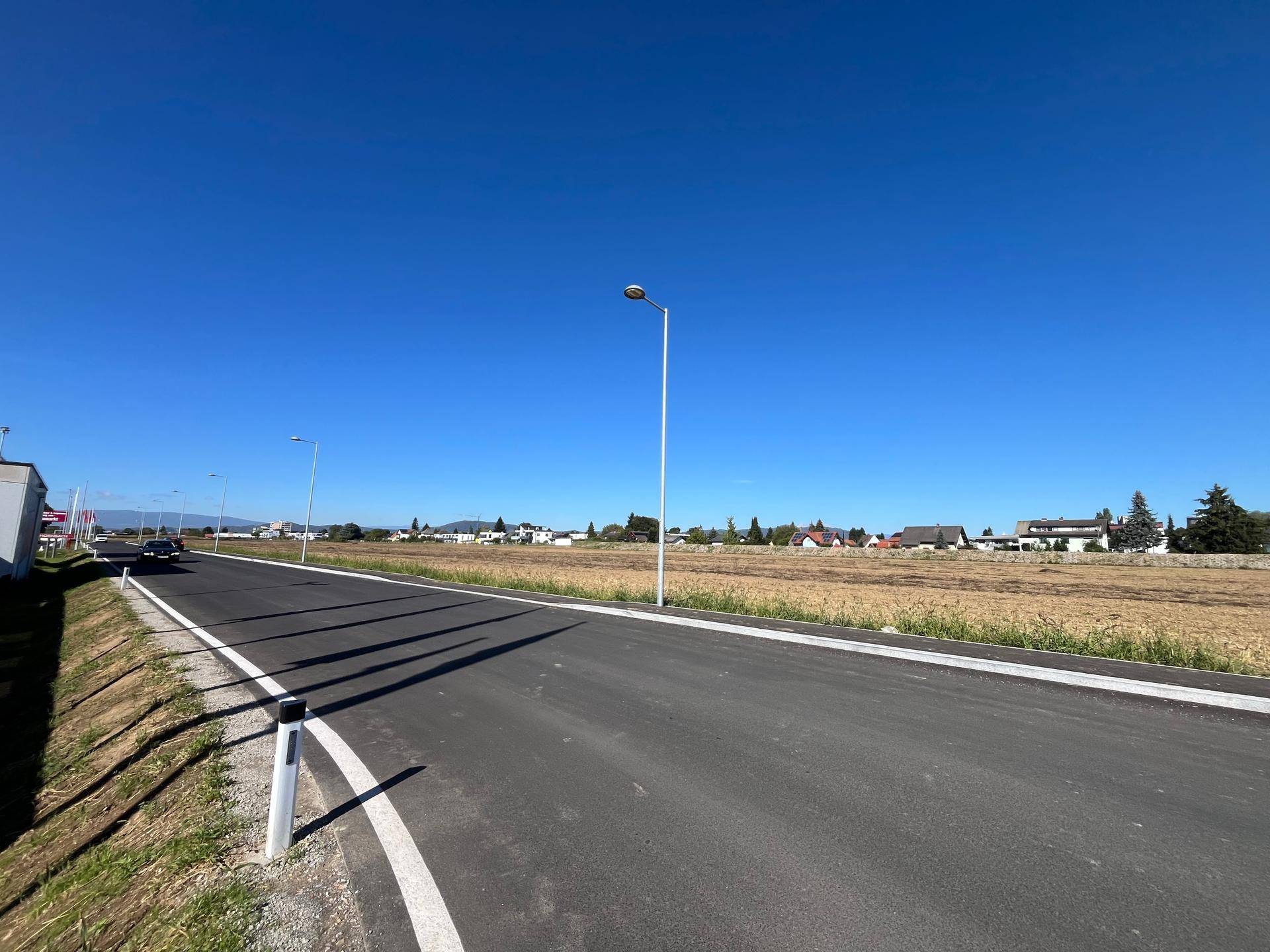 Frisch asphaltierte Straße führt entlang eines Feldes, mit einer Reihe von Häusern und Bäumen in der Ferne.