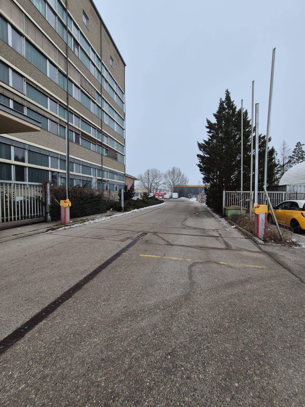 Zufahrtsstraße zu einem Gebäude mit Schranken und einem Zaun, umgeben von Bäumen.