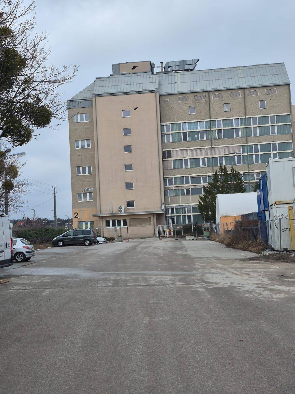 Mehrstöckiges Gebäude mit vielen Fenstern und einem Parkplatz davor unter einem grauen Himmel.