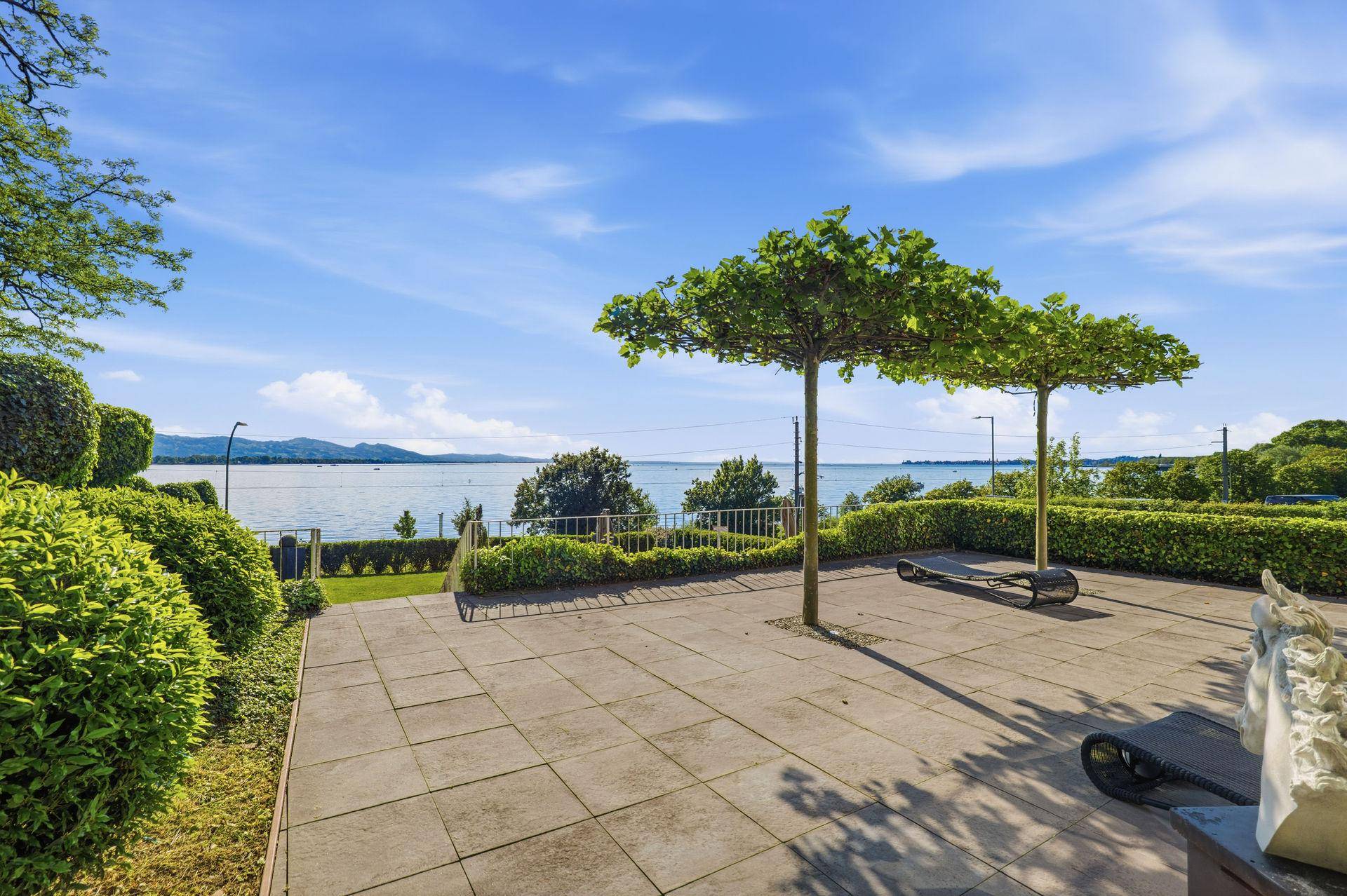 Gepflasterte Terrasse mit Liegestühlen und Hecke, die einen weiten Blick auf den See bietet.
