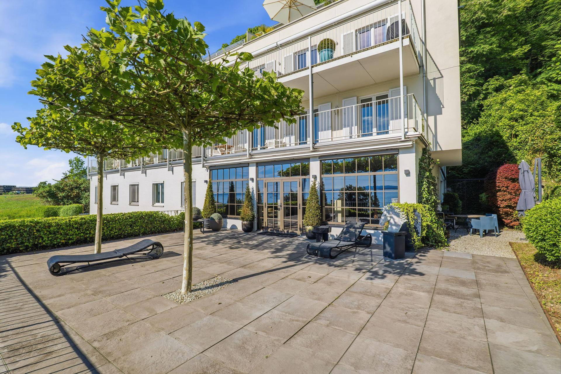 Große gepflasterte Terrasse mit Liegestühlen und Bäumen, mit Blick auf den See.