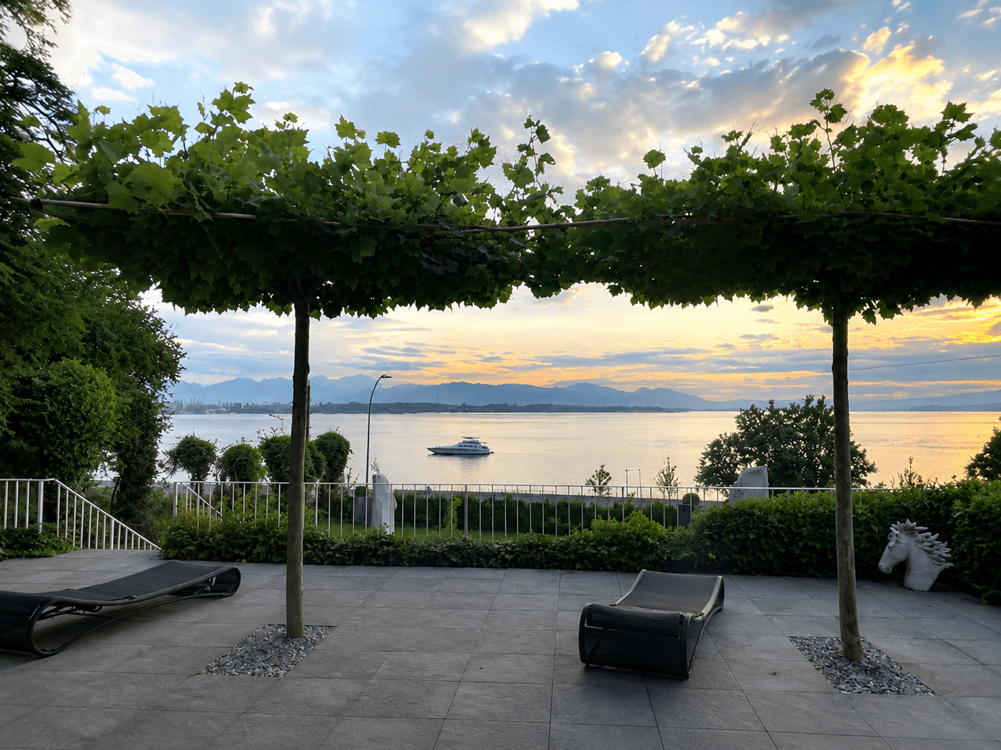 Terrasse mit Liegestühlen und Bäumen, mit malerischem Sonnenuntergangsblick über See und Berge.