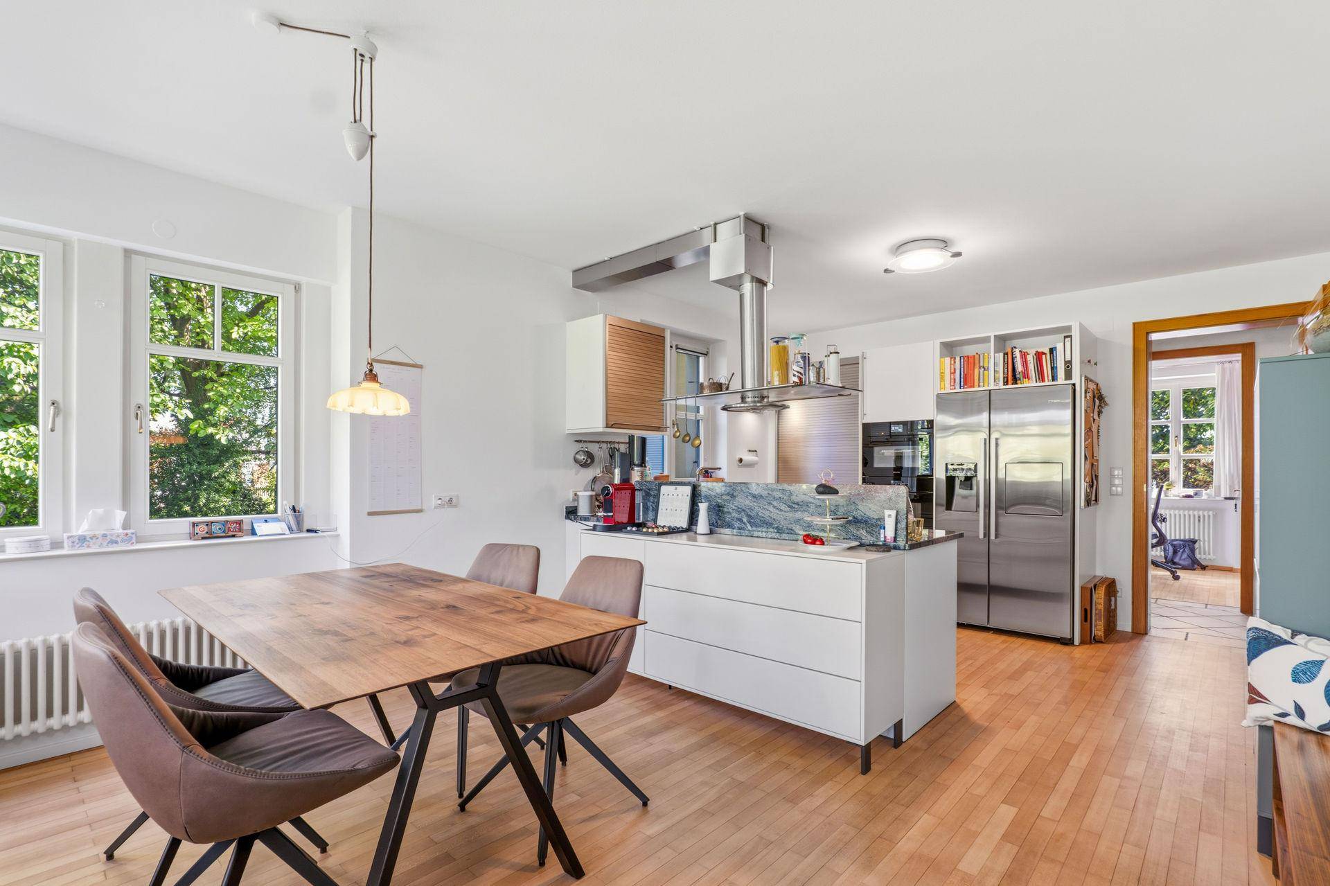 Moderne Wohnküche mit weißer Kücheninsel, Esstisch und Holzboden, hell durch große Fenster.