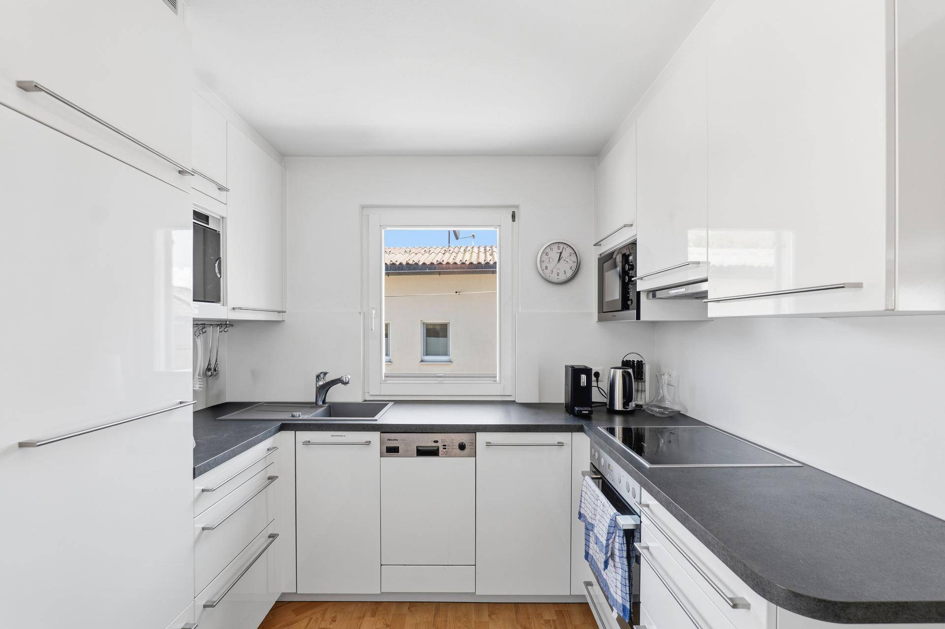 Moderne, helle Küche mit weißen Hochglanzfronten, dunklen Arbeitsplatten und Holzboden.