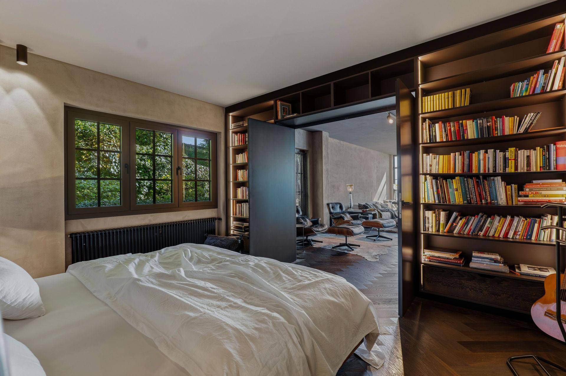 Schlafzimmer mit Bett, eingebauten Bücherregalen und Fenster, offen zum Wohnbereich.