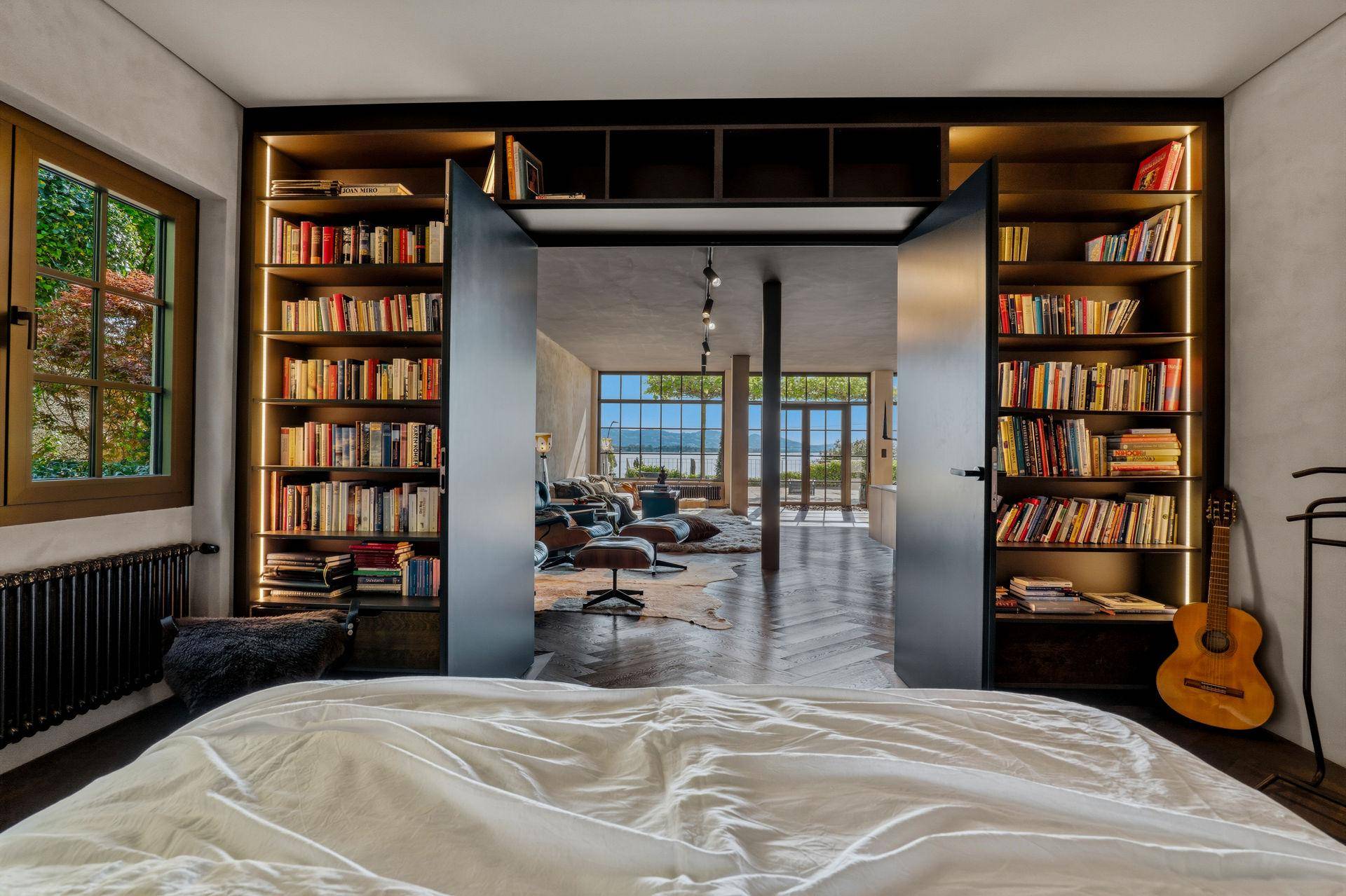 Schlafzimmer mit Bett und beleuchteten Bücherregalen, offen zum geräumigen Wohnbereich.