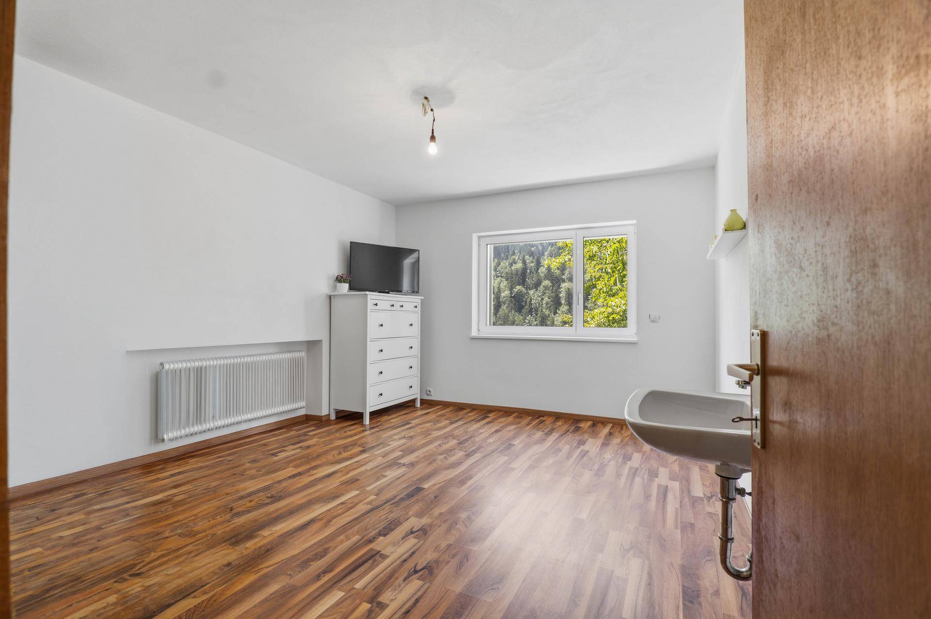 Schlafzimmer mit Holzboden, Kommode, Fernseher und Blick ins Grüne.