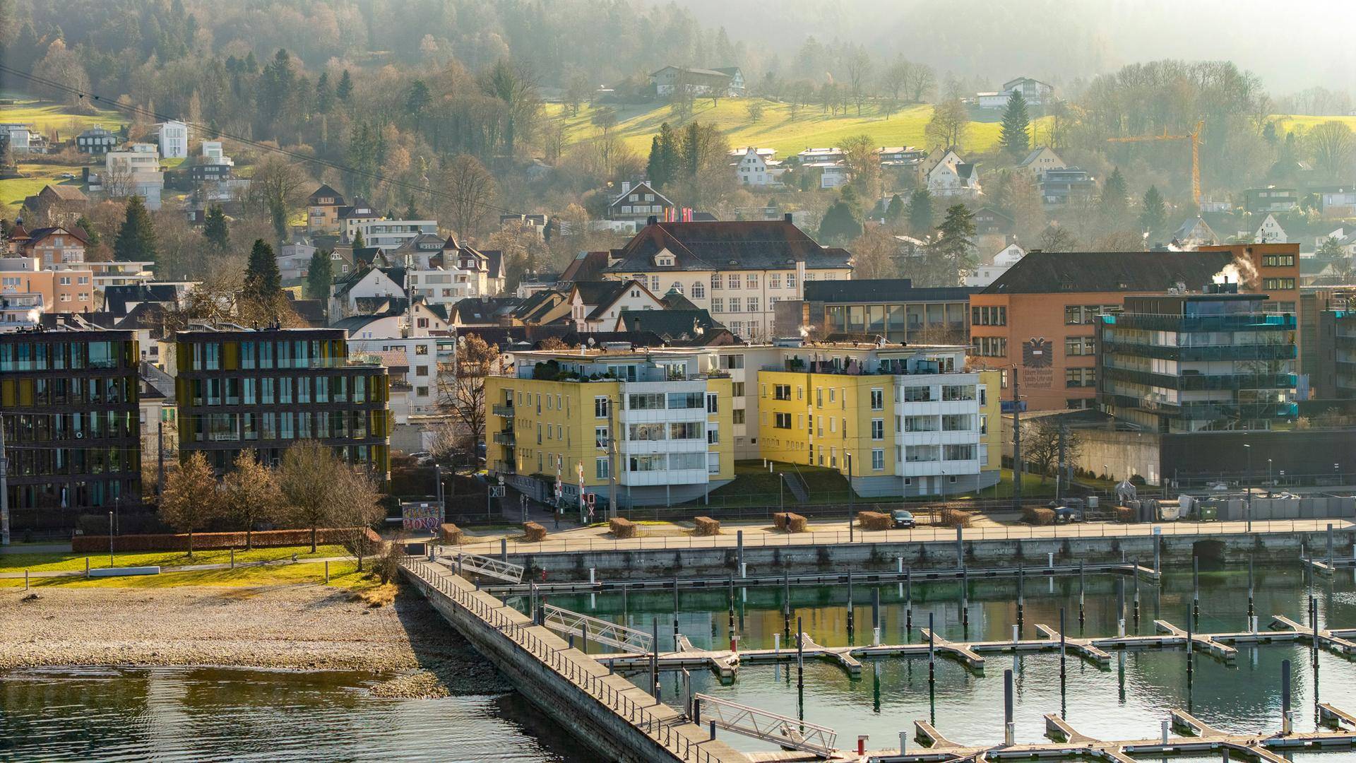 Blick auf die modernen Gebäude am Seeufer mit Yachthafen und grünen Hügeln im Hintergrund.