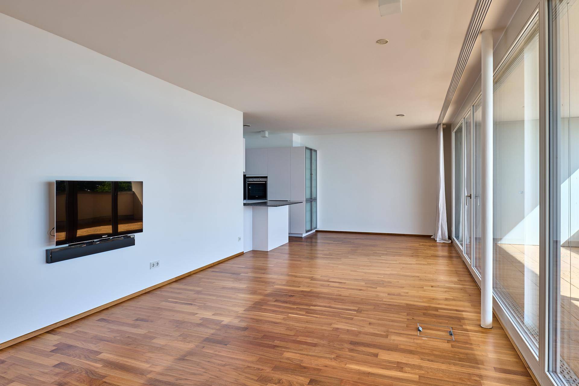 Geräumiger, heller Wohnbereich mit Holzboden und moderner Wandhalterung für einen Fernseher, offen zum Küchenbereich.