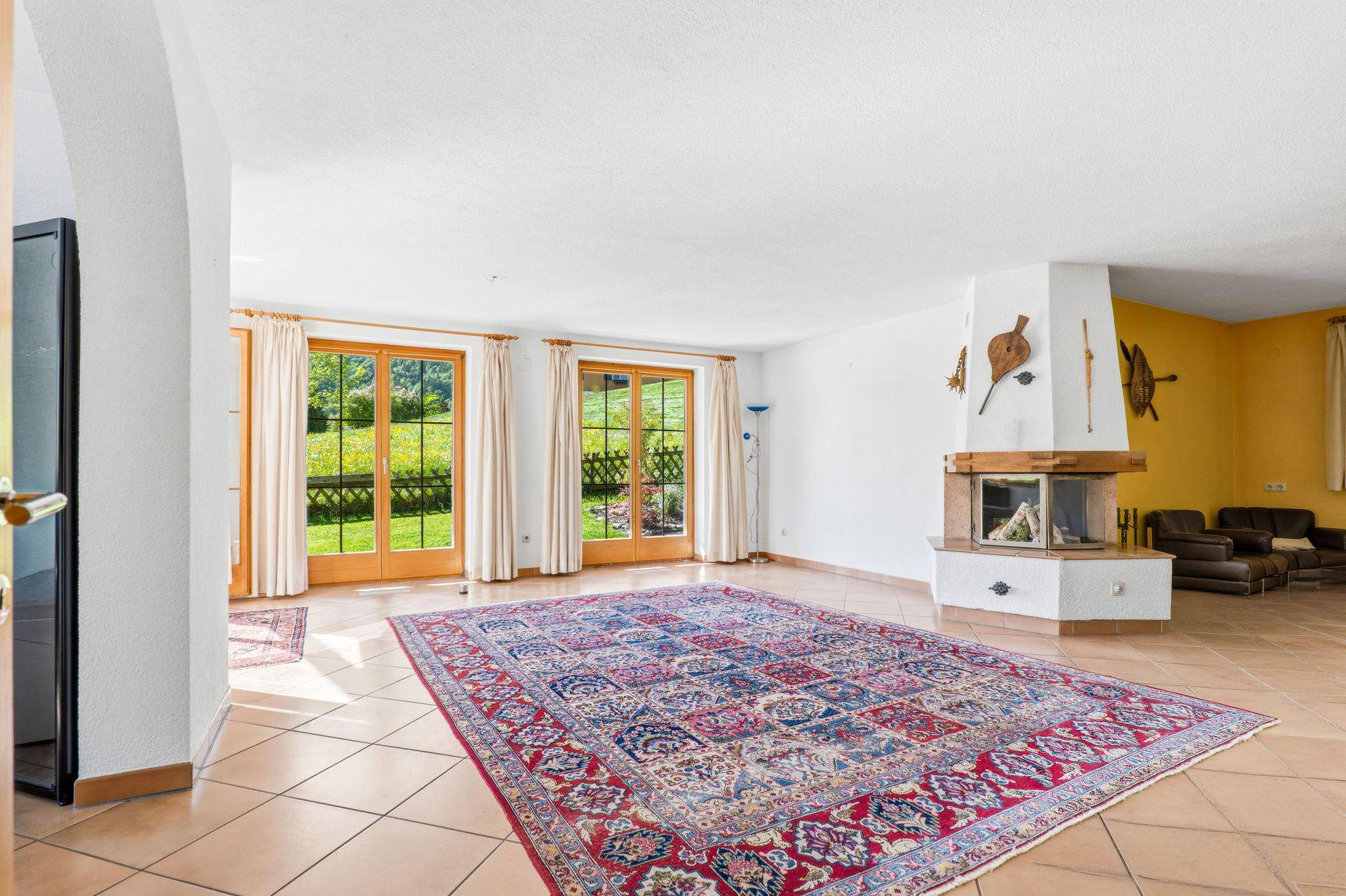 Geräumiges Wohnzimmer mit Kamin, Fliesenboden und Zugang zum Garten durch große Fenstertüren.