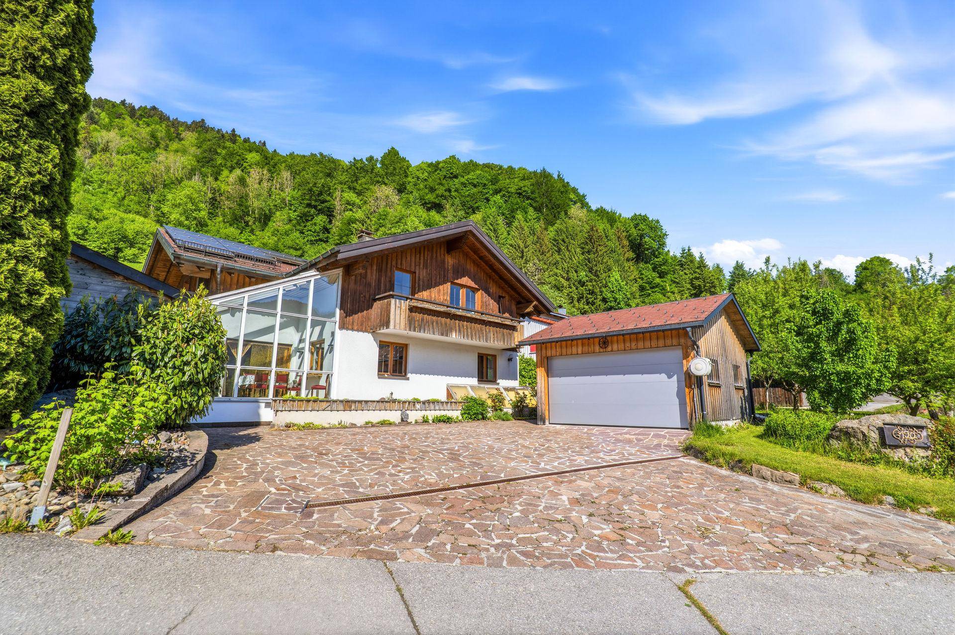 Ansicht des Hauses mit Wintergarten, gepflasterter Auffahrt und separater Garage vor grüner Bergkulisse.