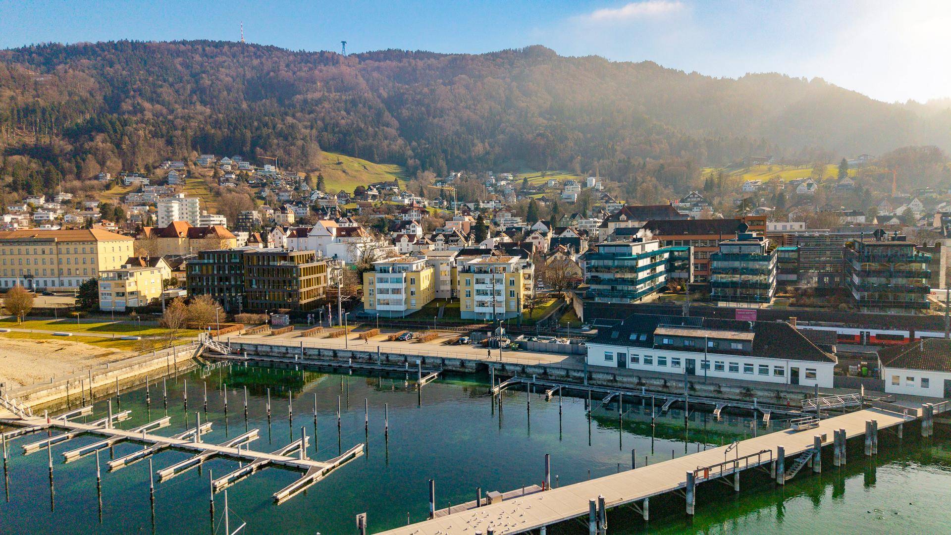 Weitläufiger Blick auf die Küstenstadt mit Yachthafen, See und bewaldeten Hügeln im Sonnenlicht.