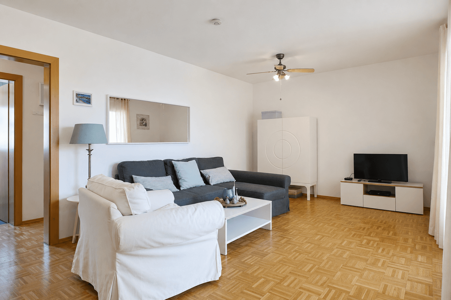 Helles Wohnzimmer mit bequemem Sofa, Parkettboden und einem Deckenventilator für angenehmes Klima.