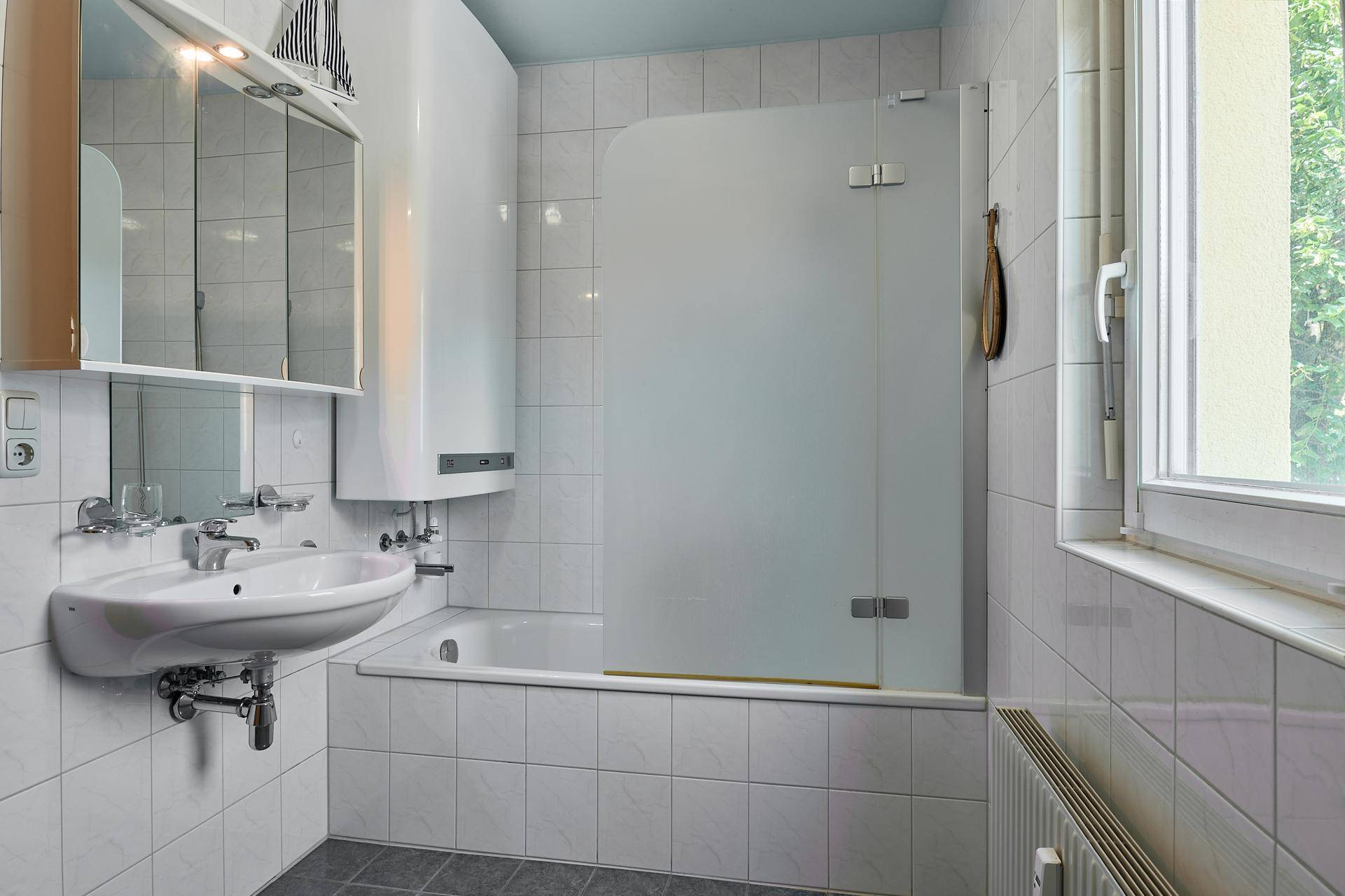 Gefliestes Badezimmer mit Badewanne, Duschwand, Waschbecken und Fenster für natürliche Belüftung.