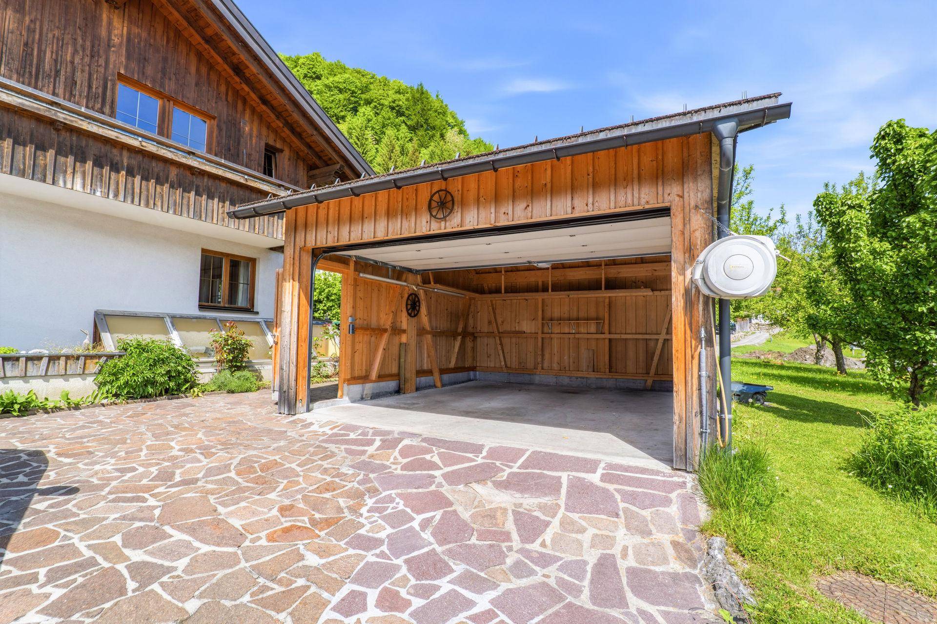Offene Holzgarage mit gepflasterter Zufahrt und angrenzendem Gartenbereich.
