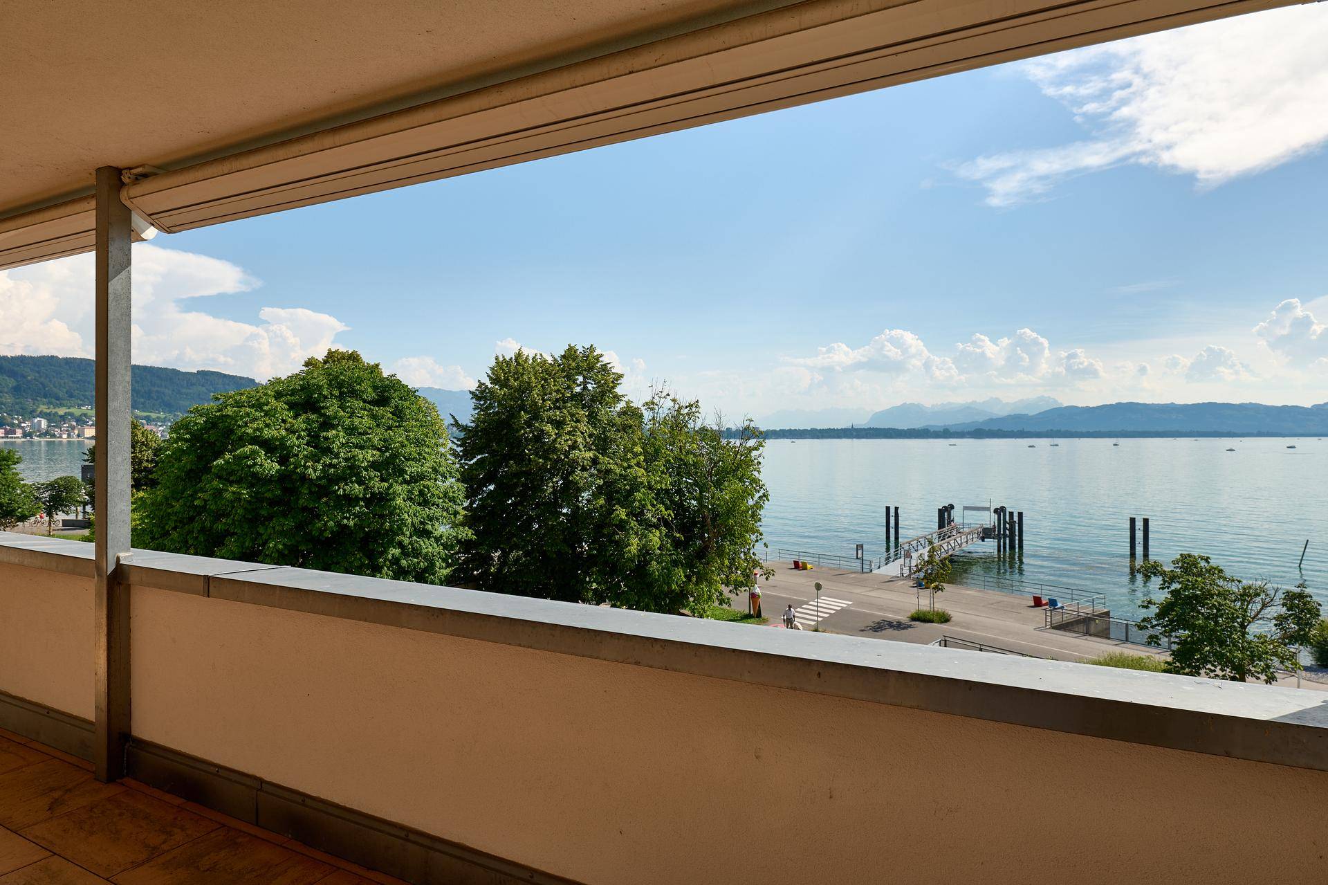 Balkon mit Blick auf den See, einen Bootssteg und üppige Bäume unter blauem Himmel.