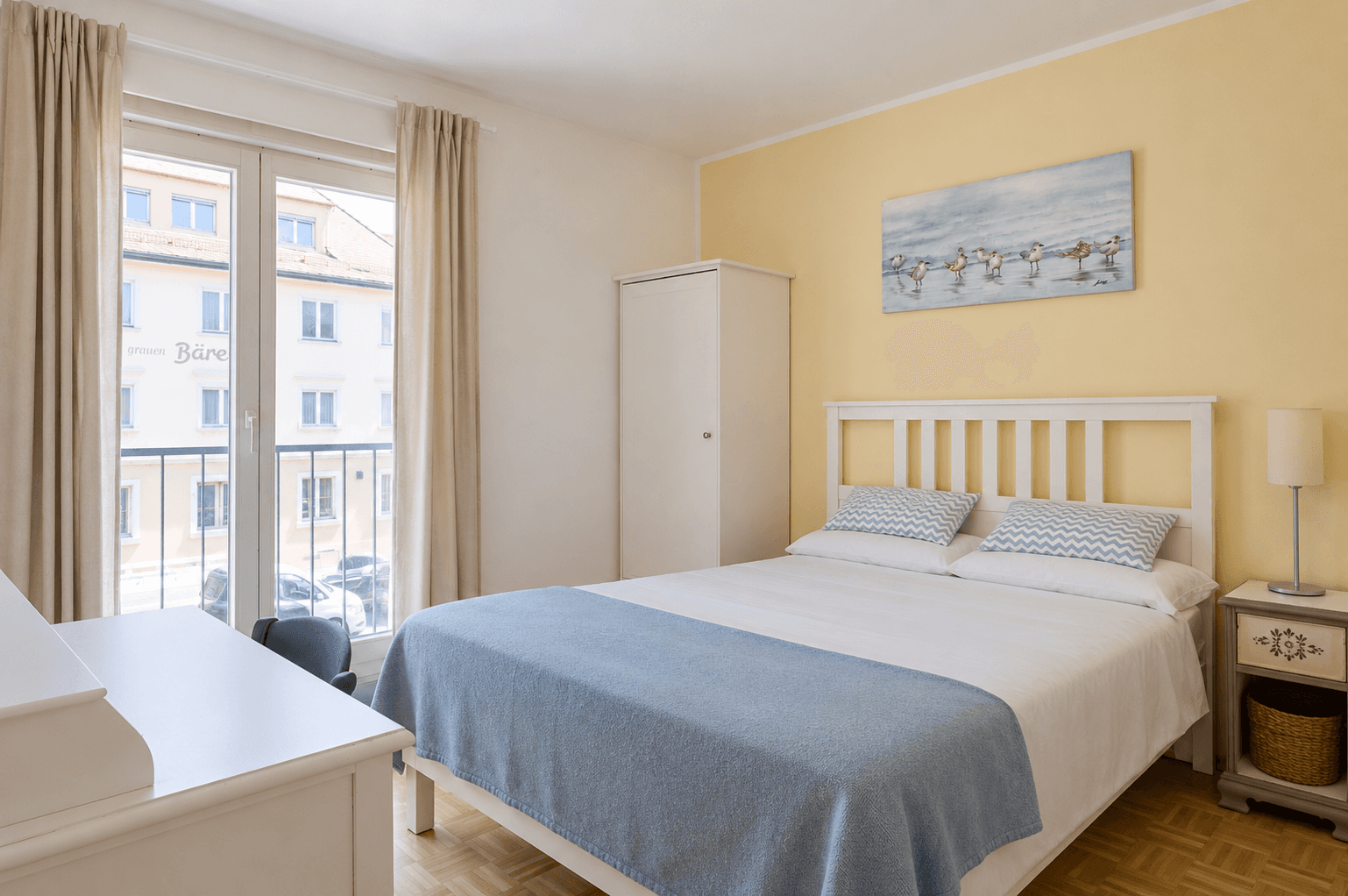 Helles Schlafzimmer mit weißem Bett, Kleiderschrank und französischem Balkon für frische Luft.