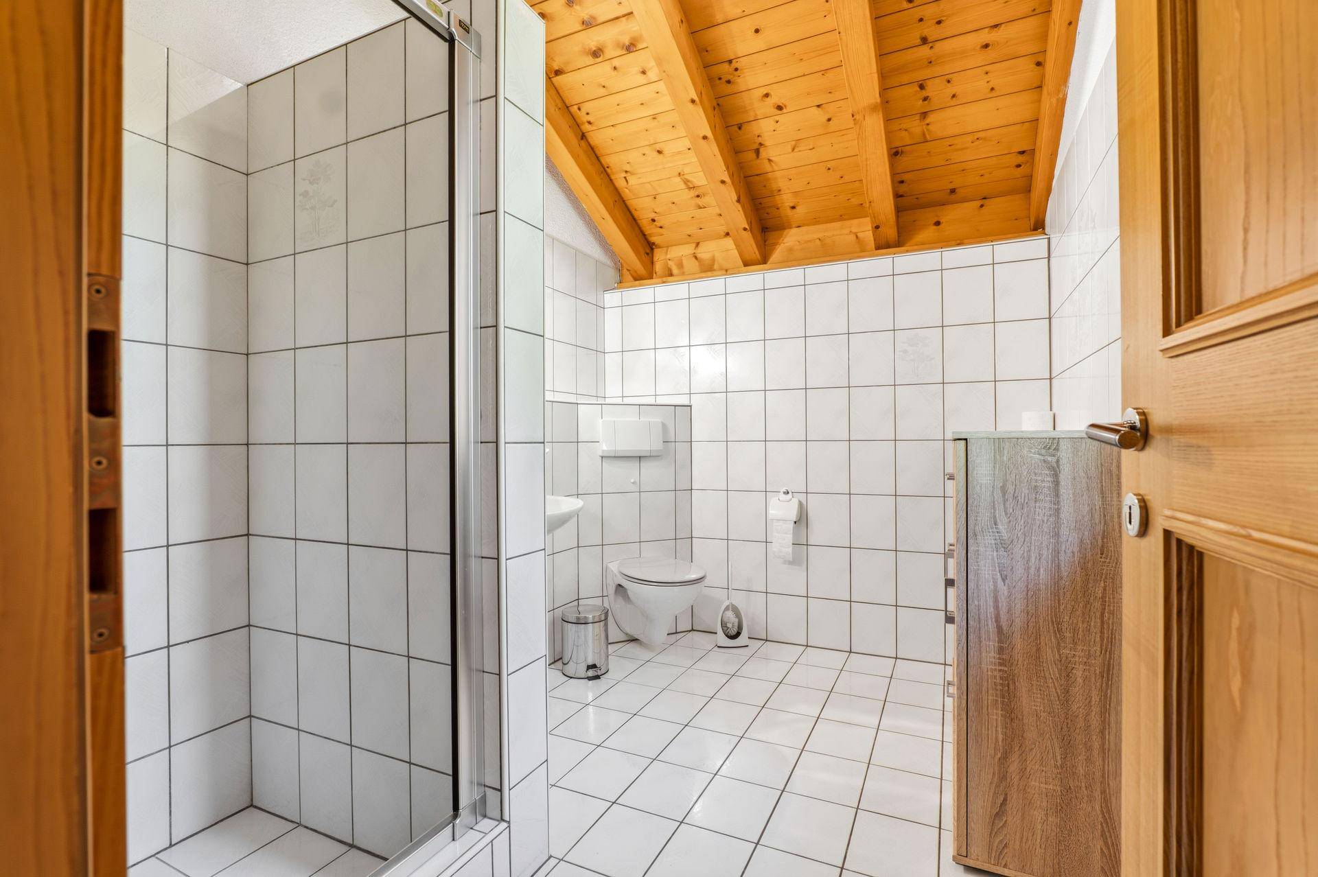 Helles Badezimmer mit Duschkabine, WC, weißen Fliesen und Holzdecke im Dachgeschoss.