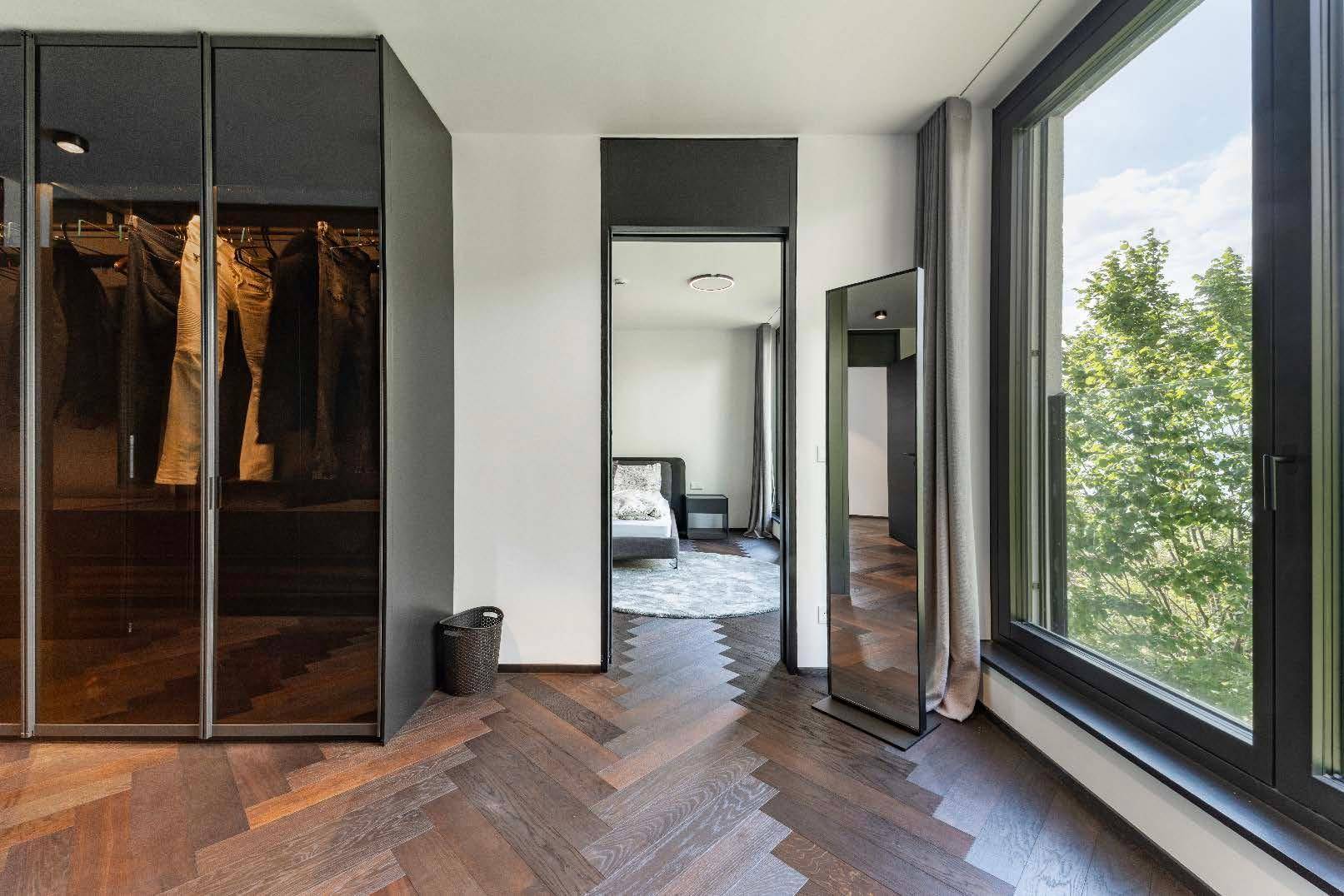 Schlafzimmerbereich mit dunklem Fischgrätparkett, raumhohen Fenstern und integriertem Kleiderschrank.