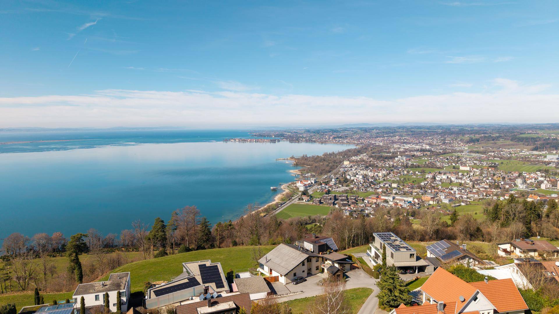 Weitläufige Luftaufnahme einer Küstenstadt am Seeufer, umgeben von grünen Hügeln unter blauem Himmel.