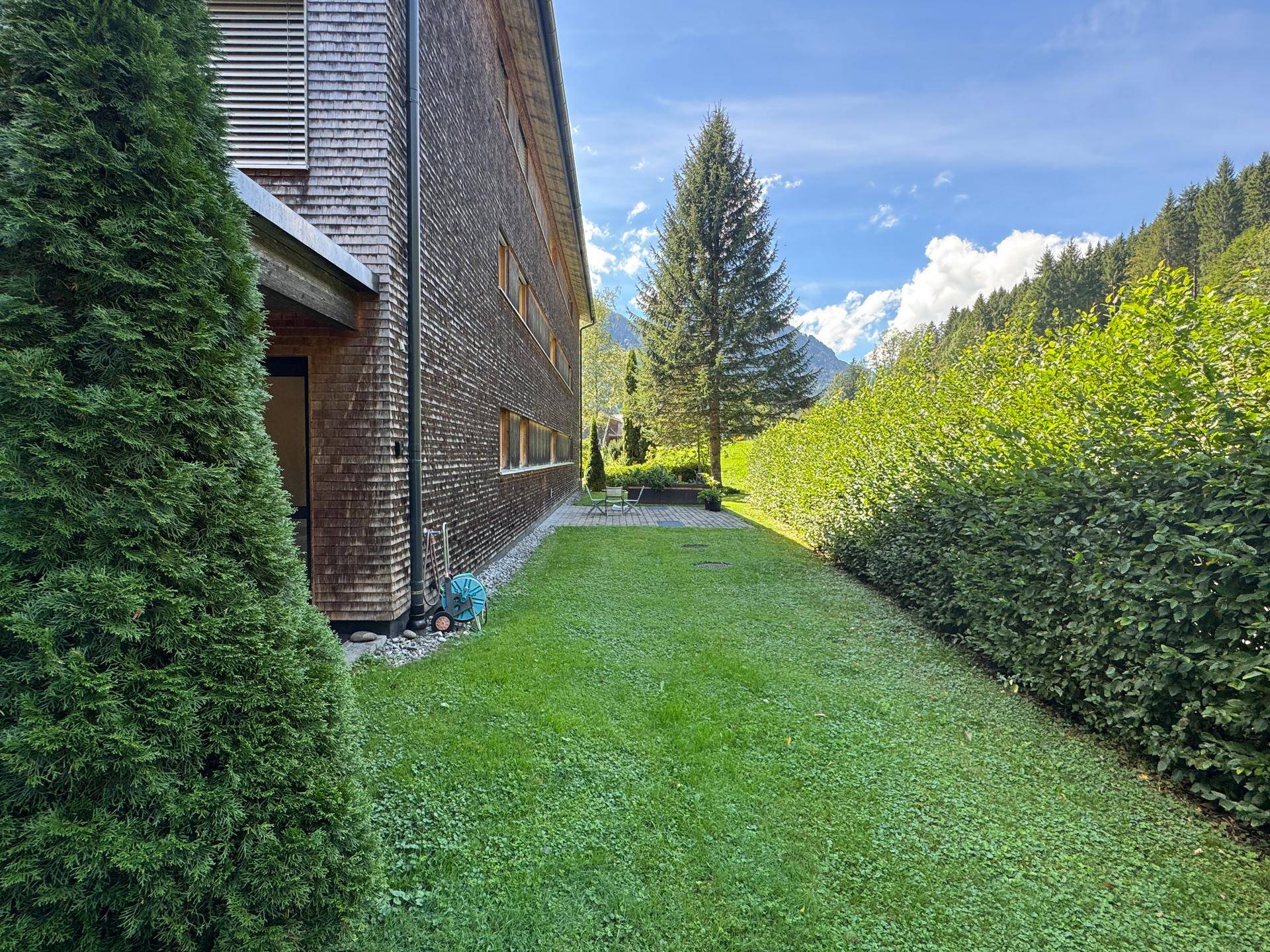 Seitlicher Gartenbereich mit gepflegtem Rasen, Hecke und Blick auf die umliegende Berglandschaft.