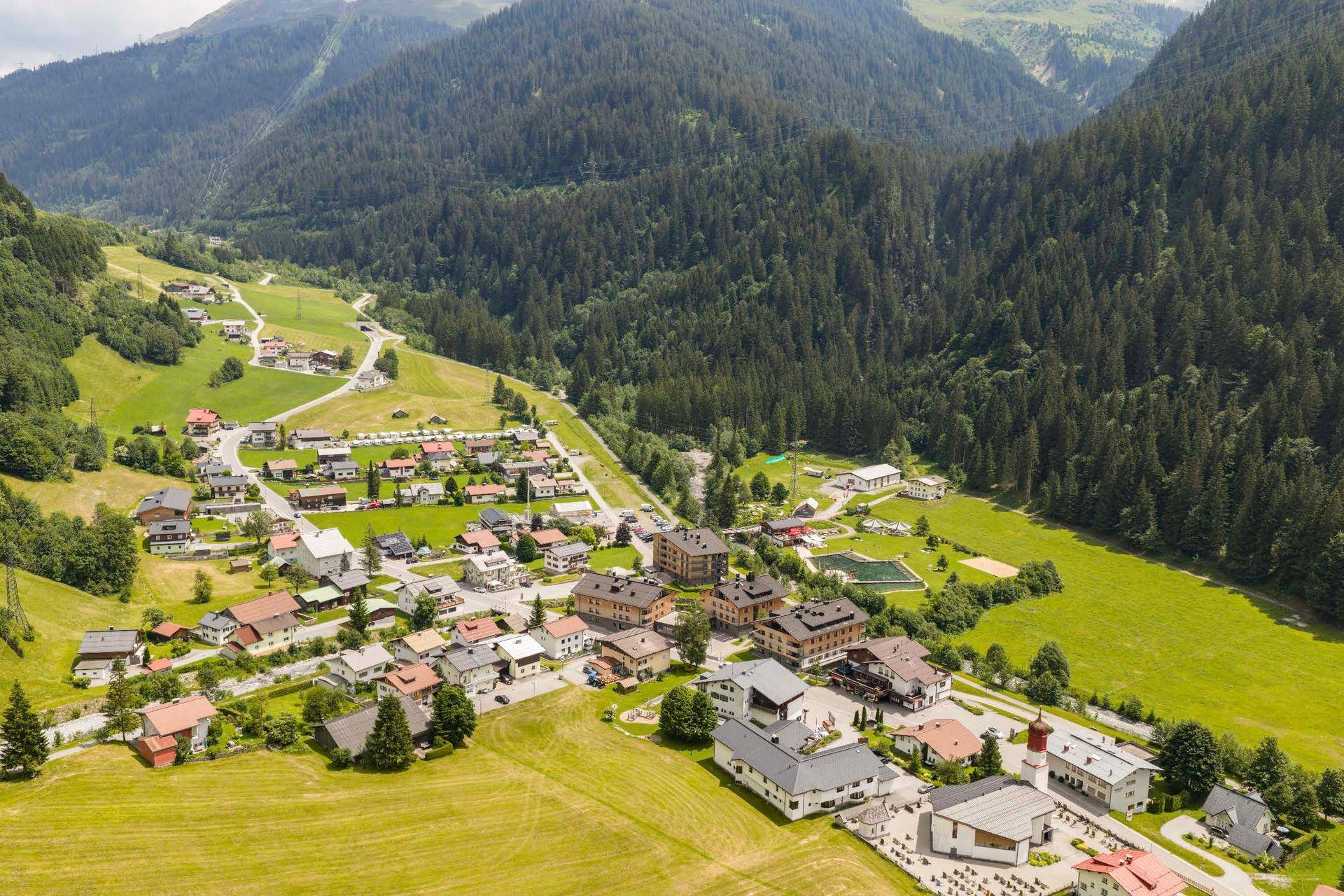 Luftaufnahme eines malerischen Dorfes in einem grünen Tal, umgeben von bewaldeten Bergen und Wiesen.