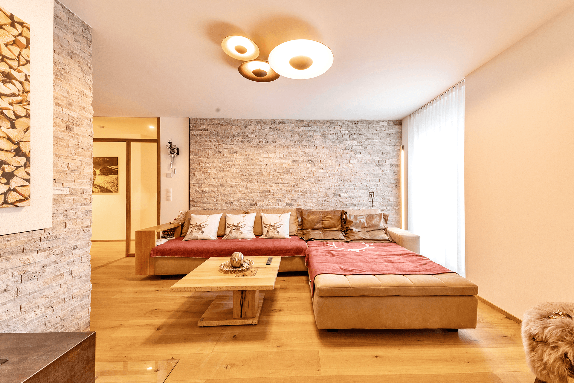 Gemütliches Wohnzimmer mit Holzboden, Steinwand und bequemer Eckcouch, beleuchtet von modernen Deckenleuchten.