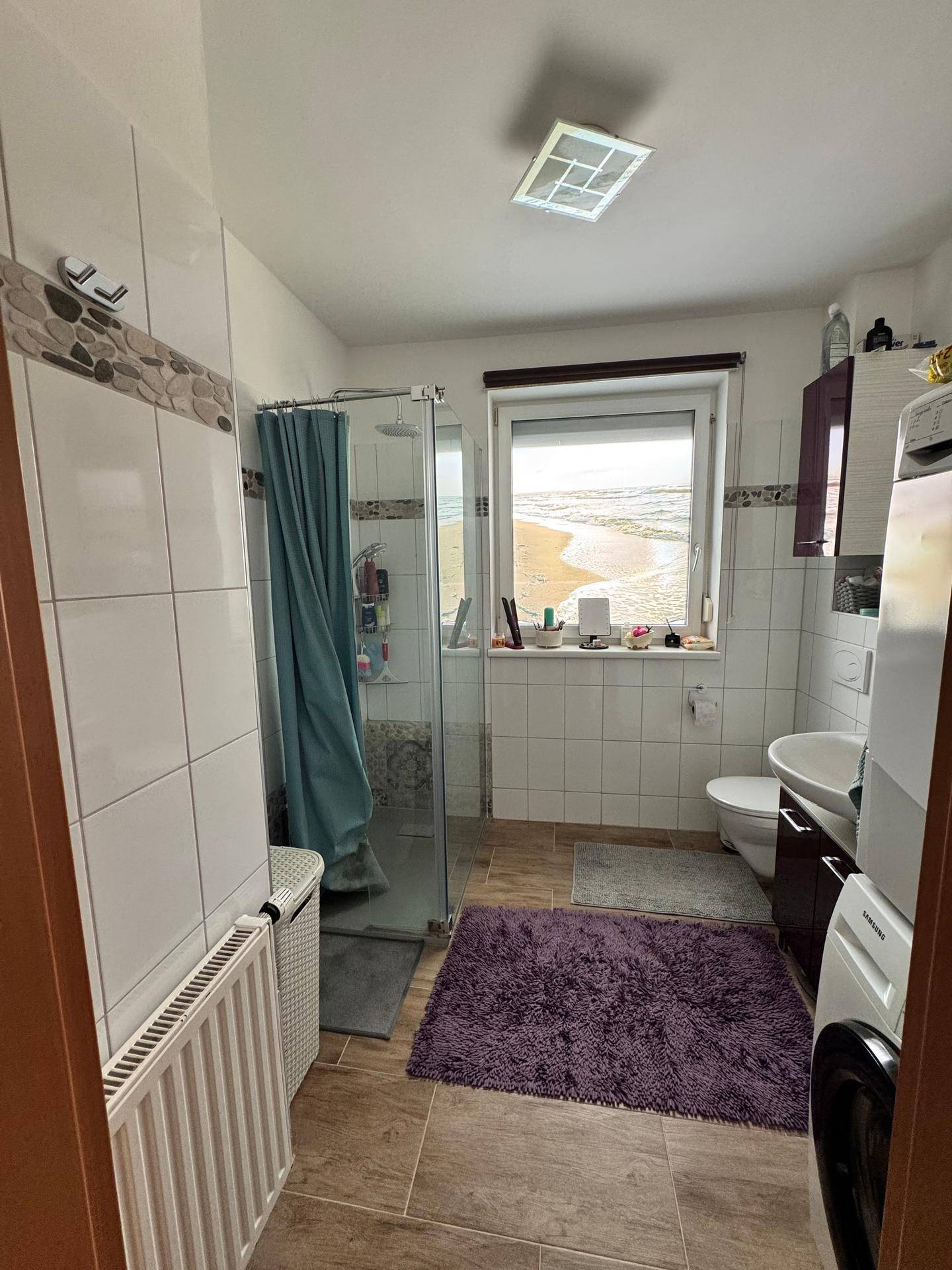 Badezimmer mit Dusche, WC, Waschbecken und Waschmaschine, mit Blick auf eine sandige Landschaft.