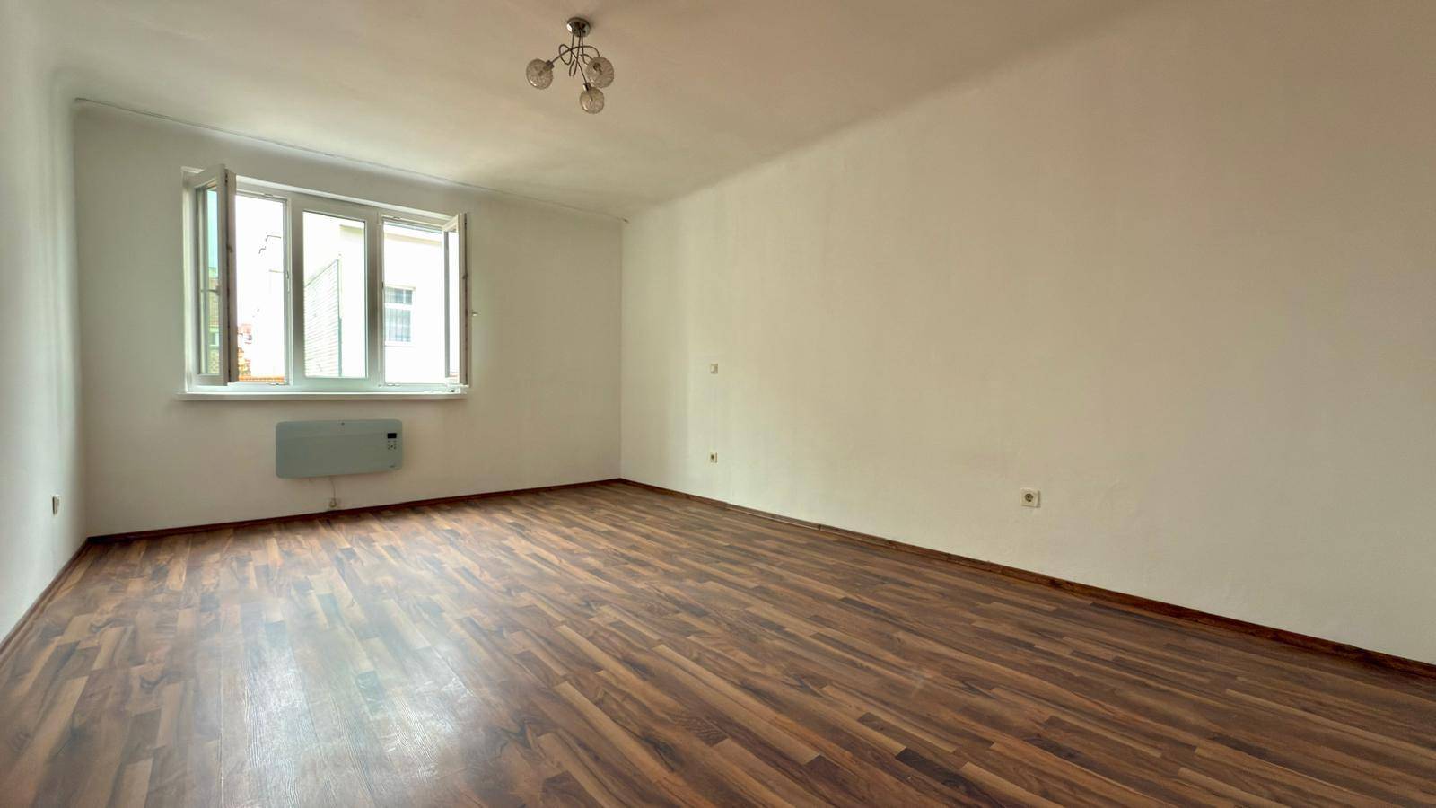 Leeres Zimmer mit Holzboden, großem Fenster und Heizkörper unter der Fensterbank.