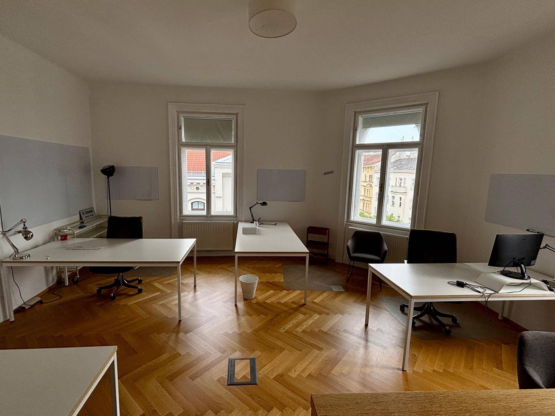 Offenes Büro mit mehreren Schreibtischen, Holzboden und zwei großen Fenstern für eine helle Arbeitsumgebung.
