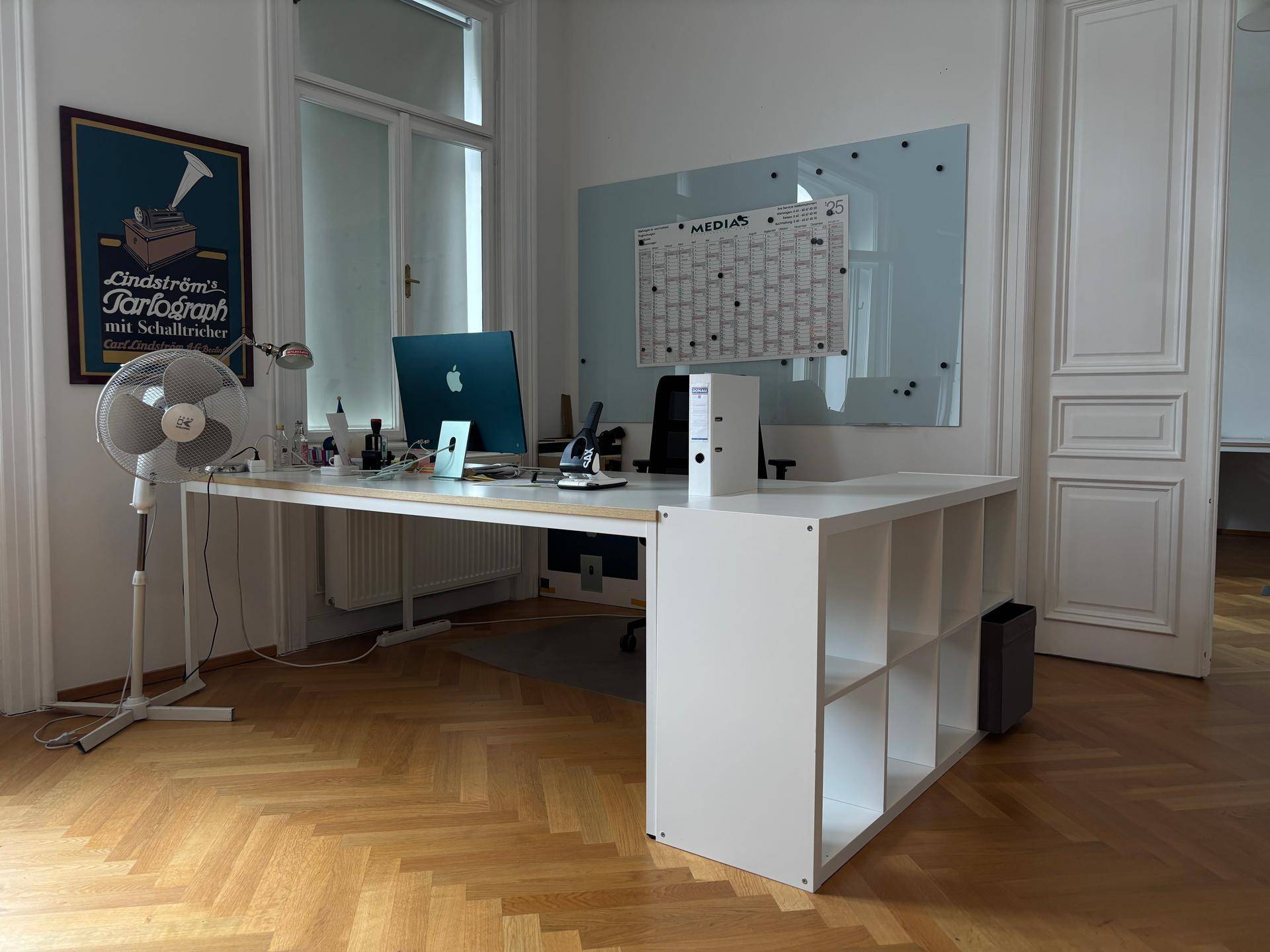 Modernes Büro mit Schreibtisch, Computer und einem Ventilator neben dem großen Fenster für angenehmes Arbeiten.