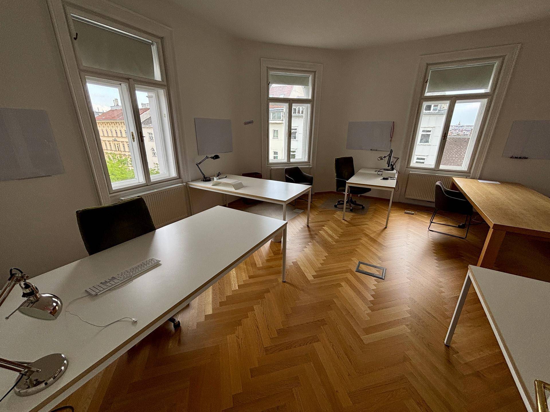 Großes Büro mit mehreren Arbeitsplätzen, Holzboden und Fenstern mit Blick auf die Stadt.