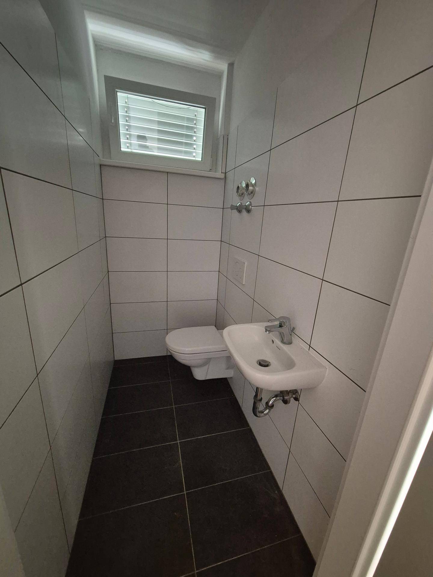 Kleines Gäste-WC mit weißen Wandfliesen, dunklem Boden und einem Fenster für Tageslicht.