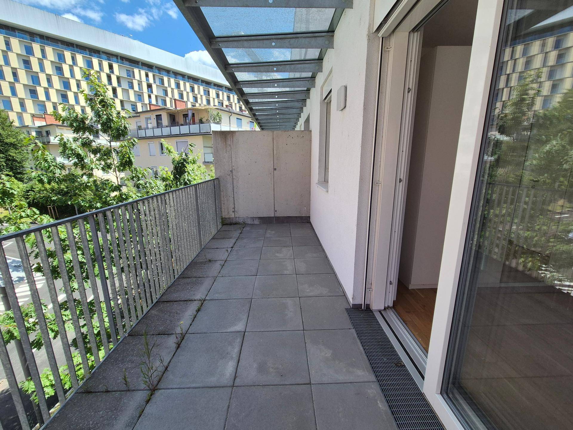 Geräumiger Balkon mit Fliesenboden und einer modernen Glasüberdachung, umgeben von Bäumen.